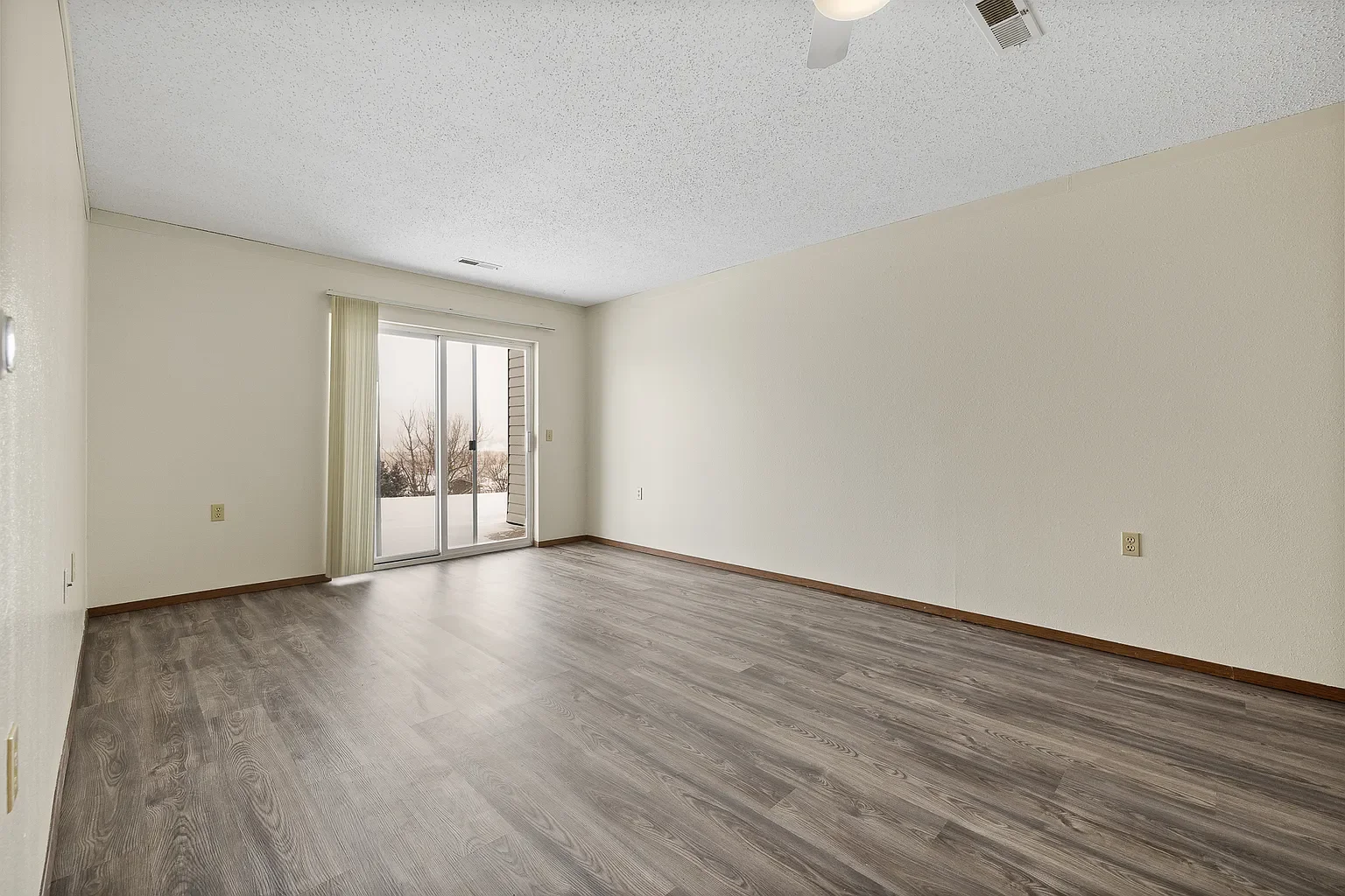 Empty living room with wooden floors, white walls, sliding glass door leading to balcony, and a ceiling fan.