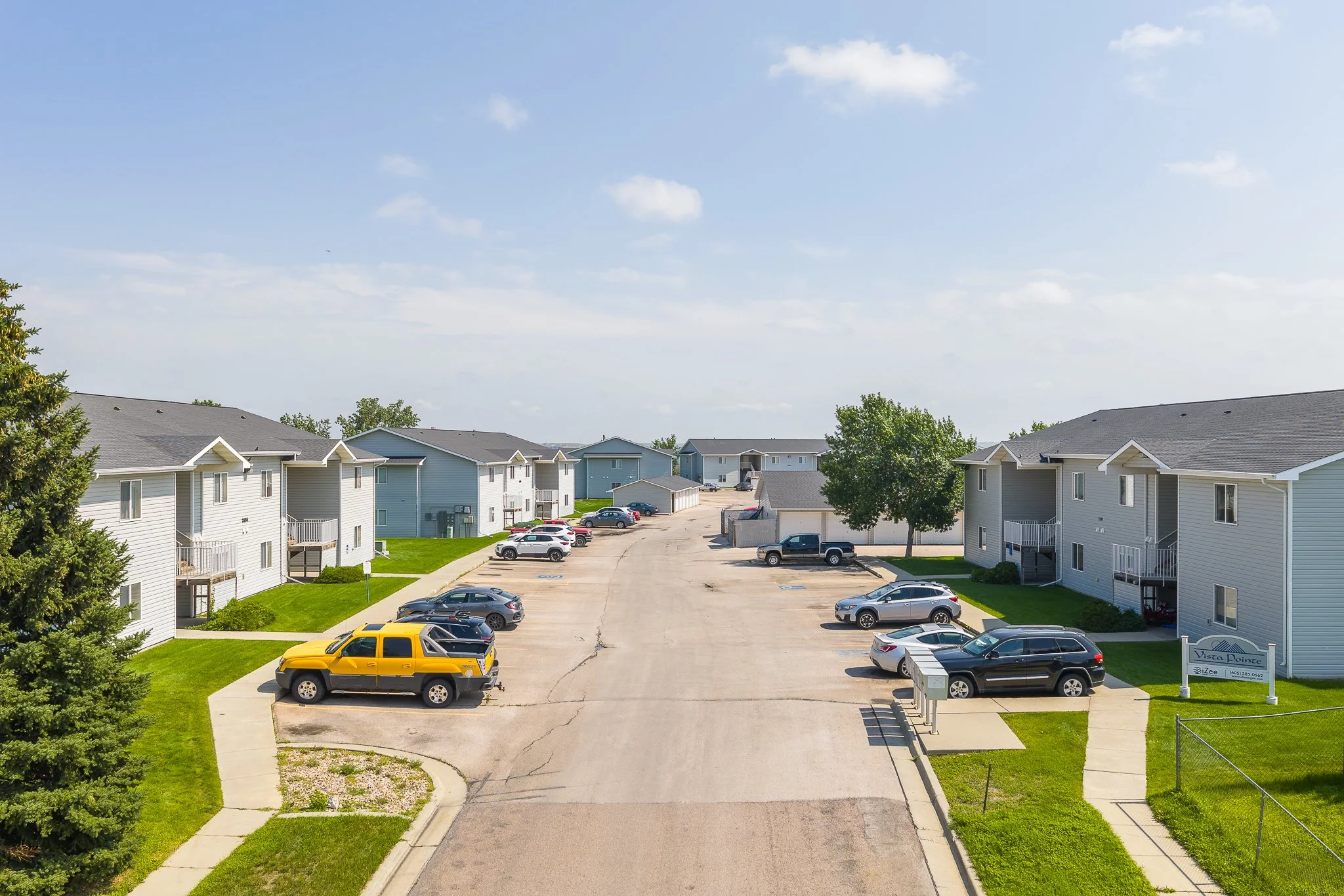 Apartment complex with multiple multi-story buildings, a parking lot with several parked cars, green lawns, trees, and a clear blue sky.