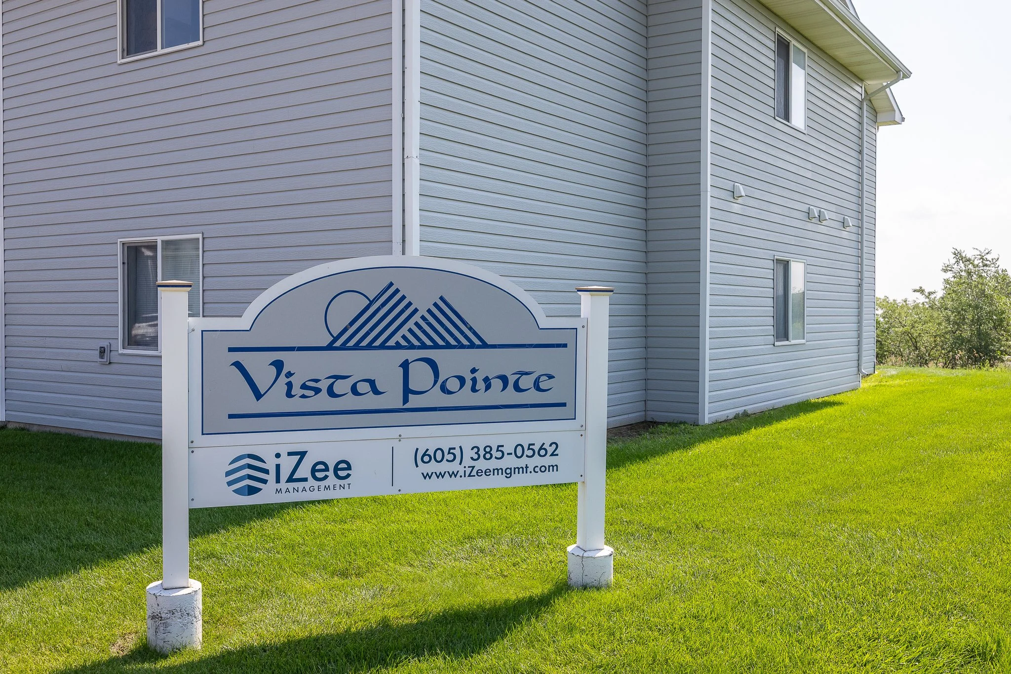 A sign in front of a light gray apartment building reads 'Vista Pointe' and has a logo with mountains and the text 'iZee Management.' The sign also displays a phone number and website address.