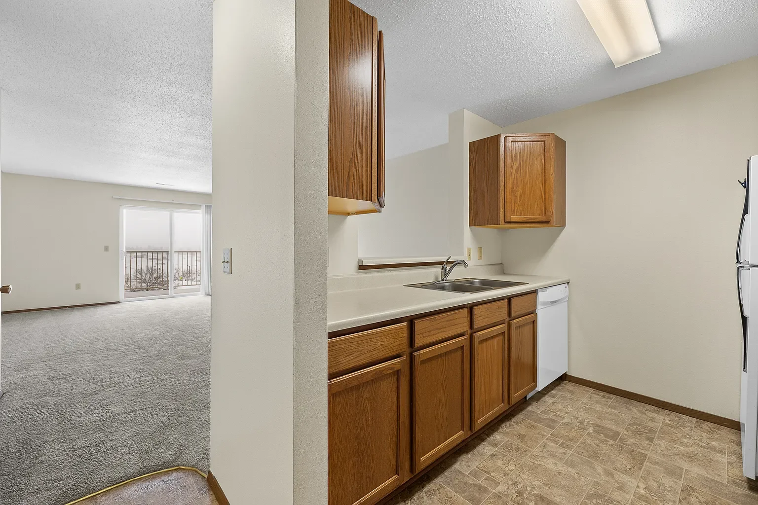 Kitchen with wooden cabinets, white countertop, and a double sink, adjacent to a living room with sliding glass door and carpeted floor.