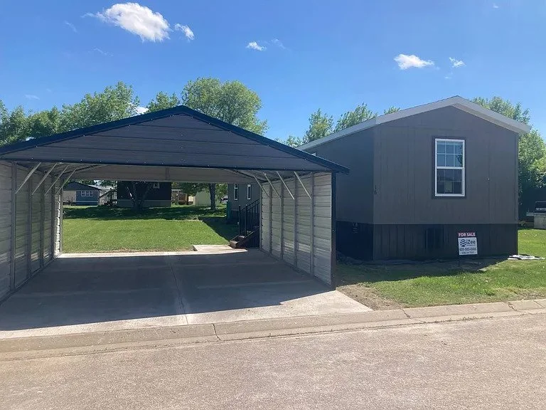 A mobile home with a metal carport on the left and a grey, two-story mobile home on the right, with a 'For Sale' sign in front, all situated on a grassy lot under a blue sky with a few clouds.