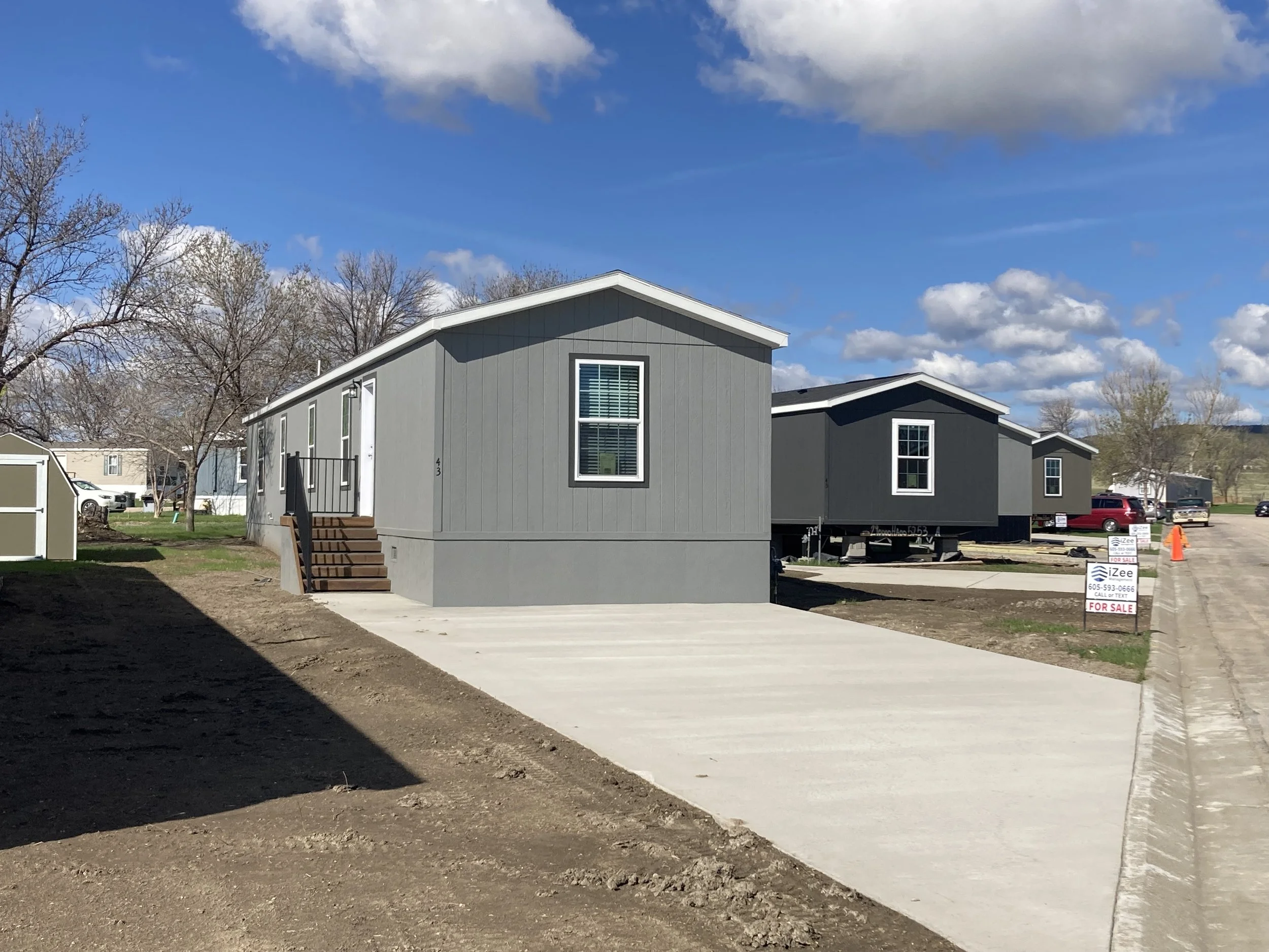 New gray manufactured homes on a lot with a concrete driveway and a blue sky with some clouds.