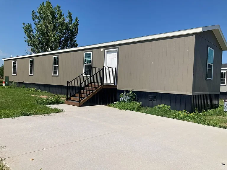 A beige manufactured home with multiple windows, a small black staircase with railings leading to a white door, and a concrete driveway in front, set against a clear blue sky.