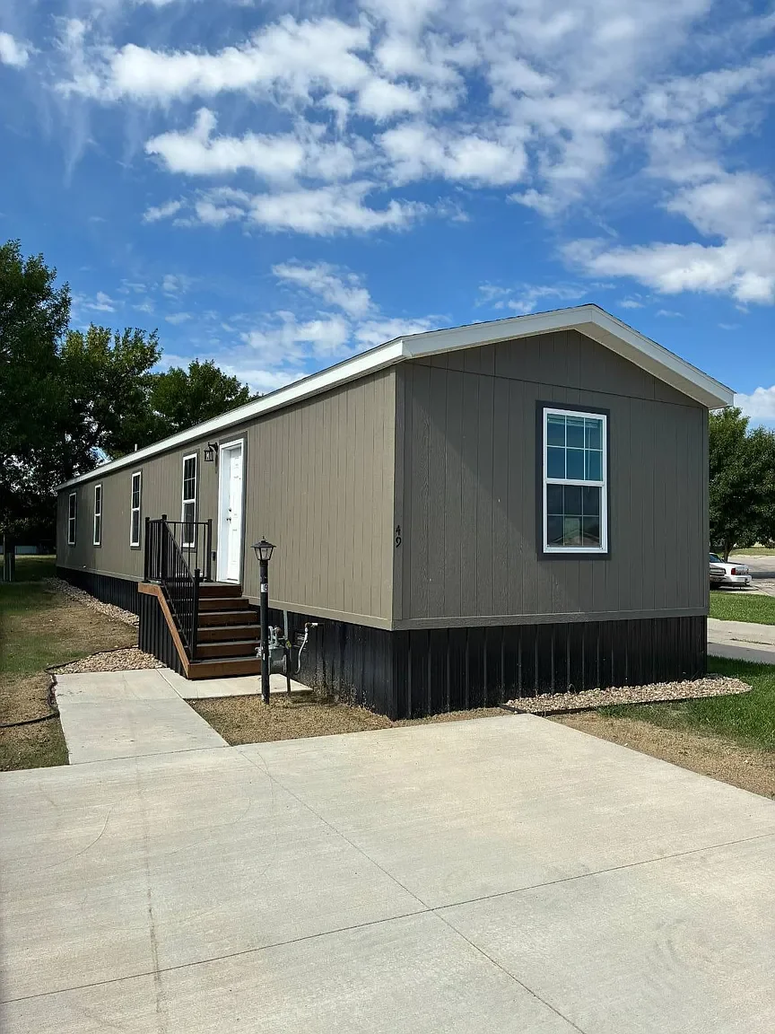 A beige manufactured home with black skirting, white door, and a small wooden staircase, situated on a concrete driveway with a landscape of grass, trees, and a blue sky with scattered clouds.