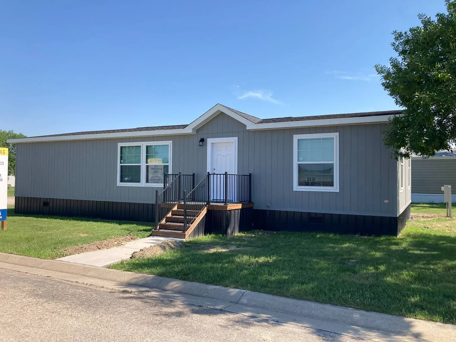 New gray manufactured home with black steps leading to a white front door, located on a green lawn under a clear blue sky.