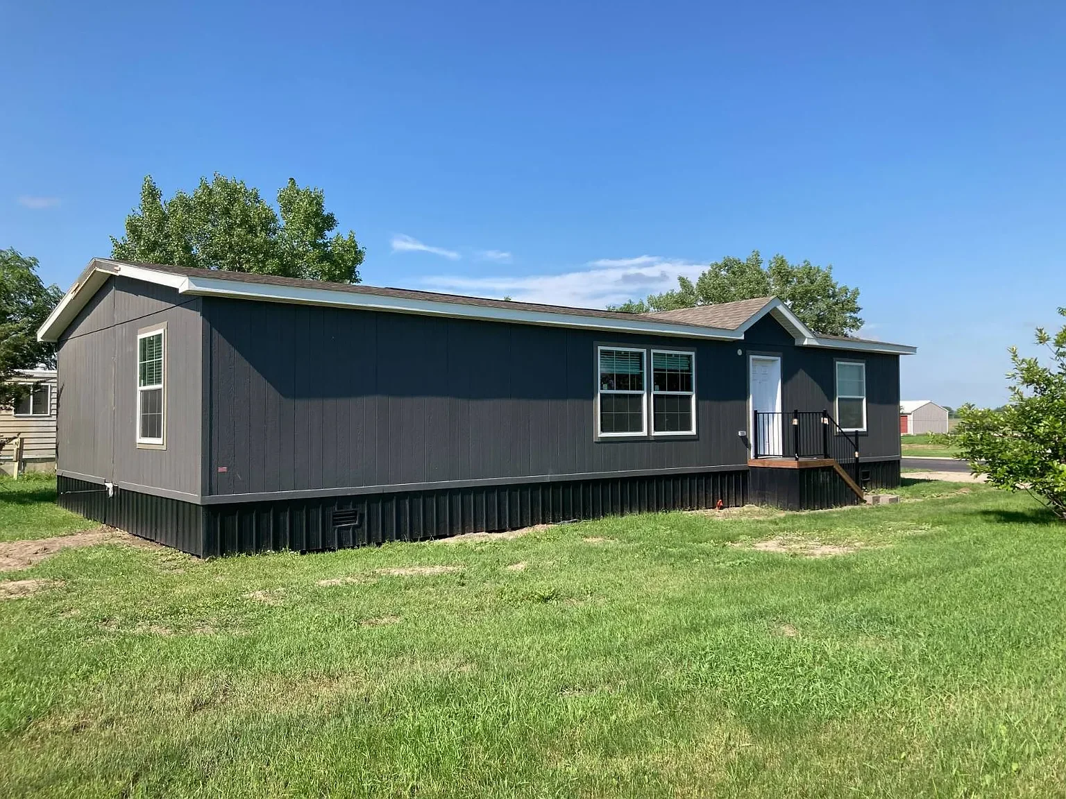 A newly constructed, single-story, dark gray modular home with white window frames, set on a green grassy lot under a bright blue sky.