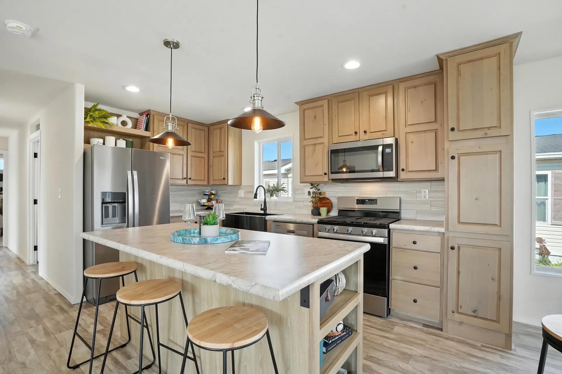 Modern kitchen with light wood cabinets, stainless steel appliances, a marble-top island with three wooden stools, pendant lighting, a window above the sink, and decorative plants.