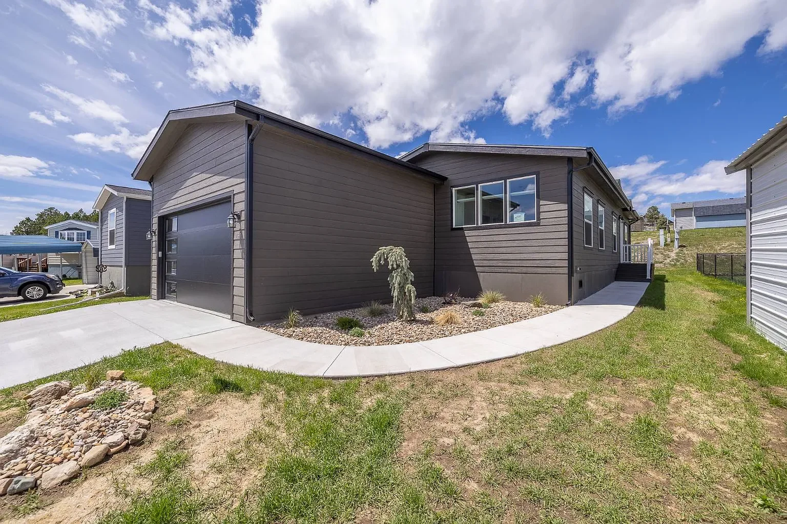 Modern single-story house with dark gray siding, attached garage, and landscaped yard under a partly cloudy sky.