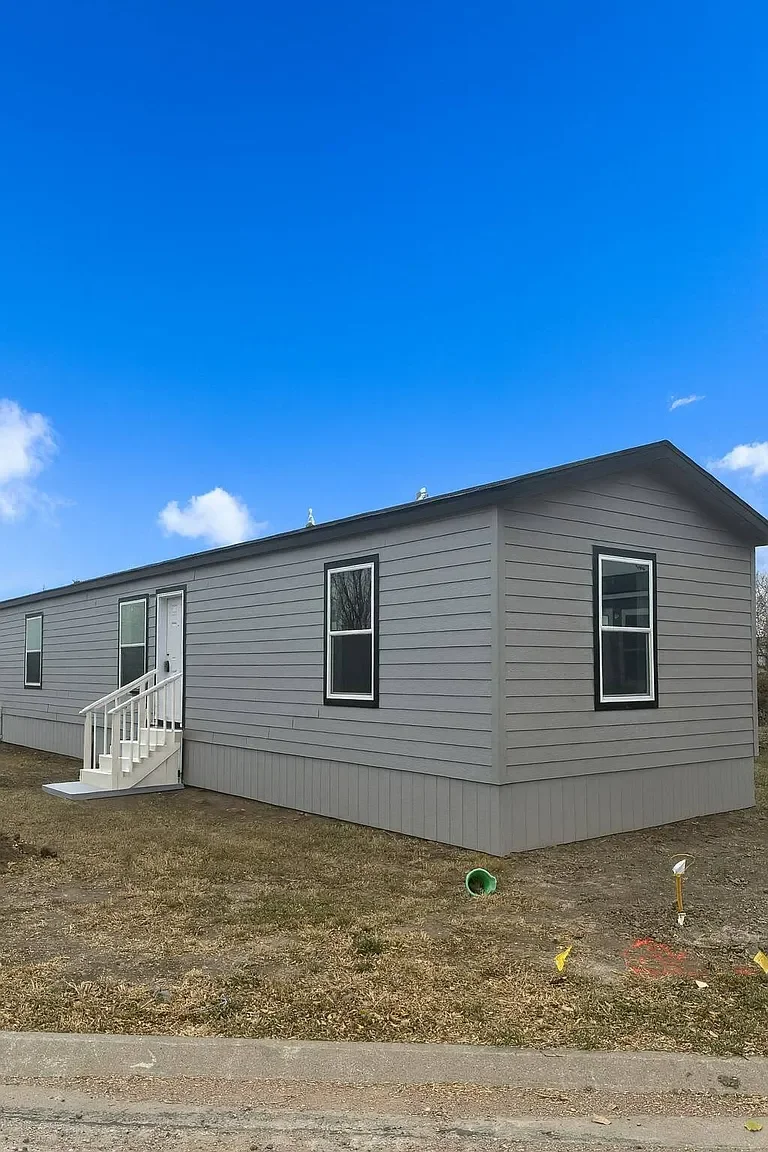 Newly constructed grey manufactured home with white steps leading to the door, sitting on a lot with patchy grass and construction markers, under a bright blue sky with a few clouds.