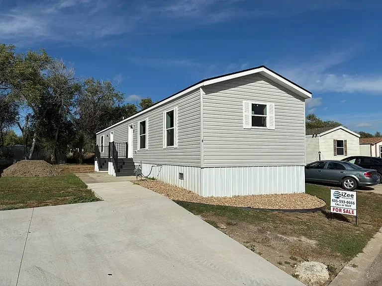 White manufactured home with steps leading to the front door, located on a concrete driveway, with a 'For Sale' sign in the yard, cars parked nearby, and a partly cloudy sky overhead.
