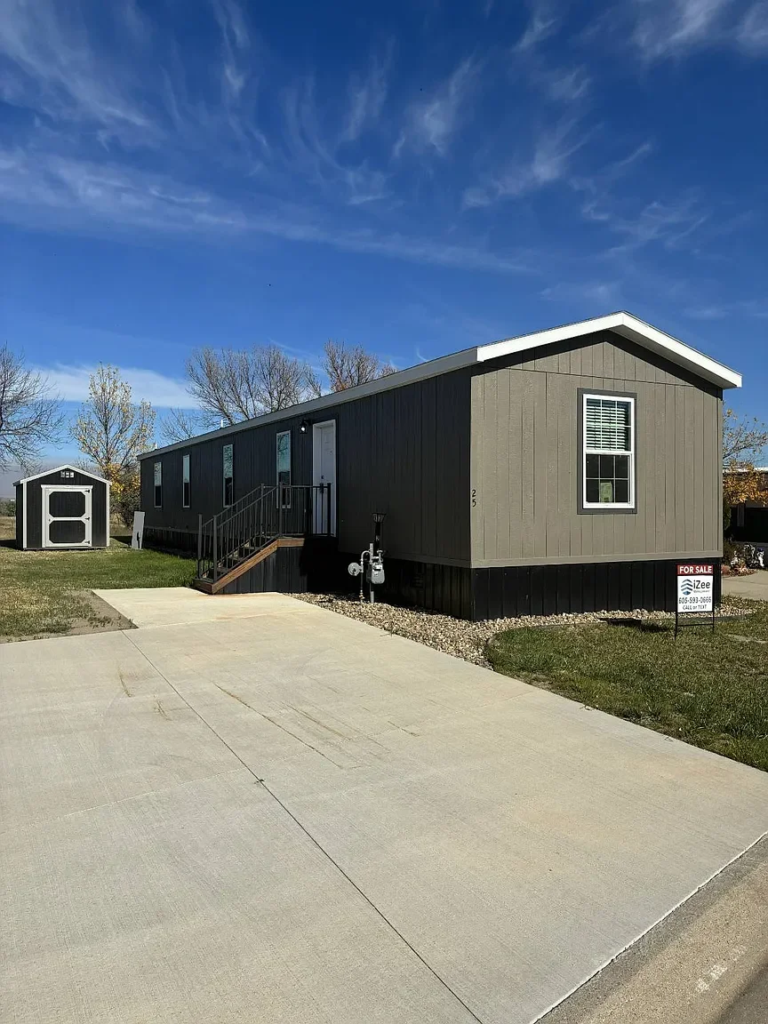 A gray manufactured home with white trim sitting on a yard with a concrete driveway, small stairs leading to the front door, and a small shed in the background, under a partly cloudy blue sky.