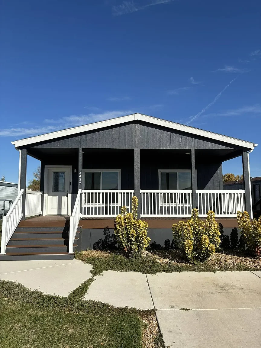 Front view of a black house with a white railing porch, stairs, and landscaped bushes, under a clear blue sky.