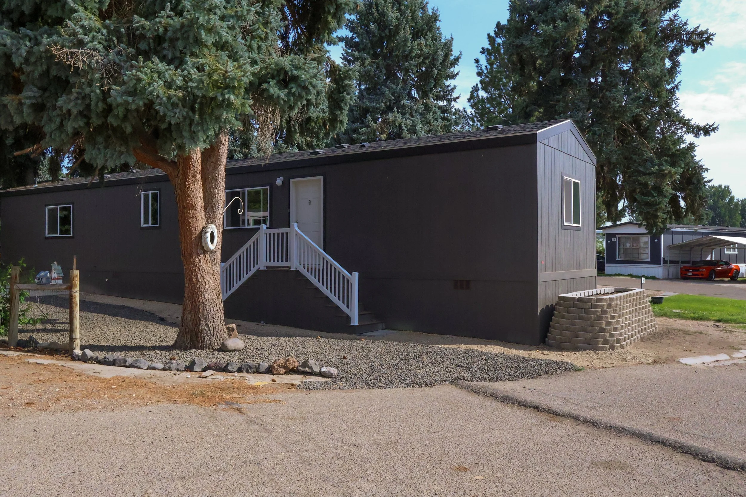 A black manufactured home with white stairs leading to a white front door, situated behind a large evergreen tree. Several windows are visible, and there is a small brick planter in front of the home. A gravel area surrounds the tree, and neighboring manufactured homes and cars are visible in the background.