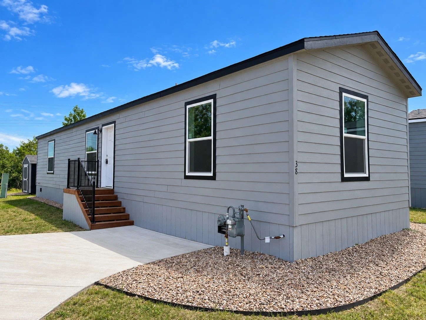 A gray manufactured home with white trim, four windows, and a small white staircase leading to the front door, set against a bright blue sky with a few clouds.