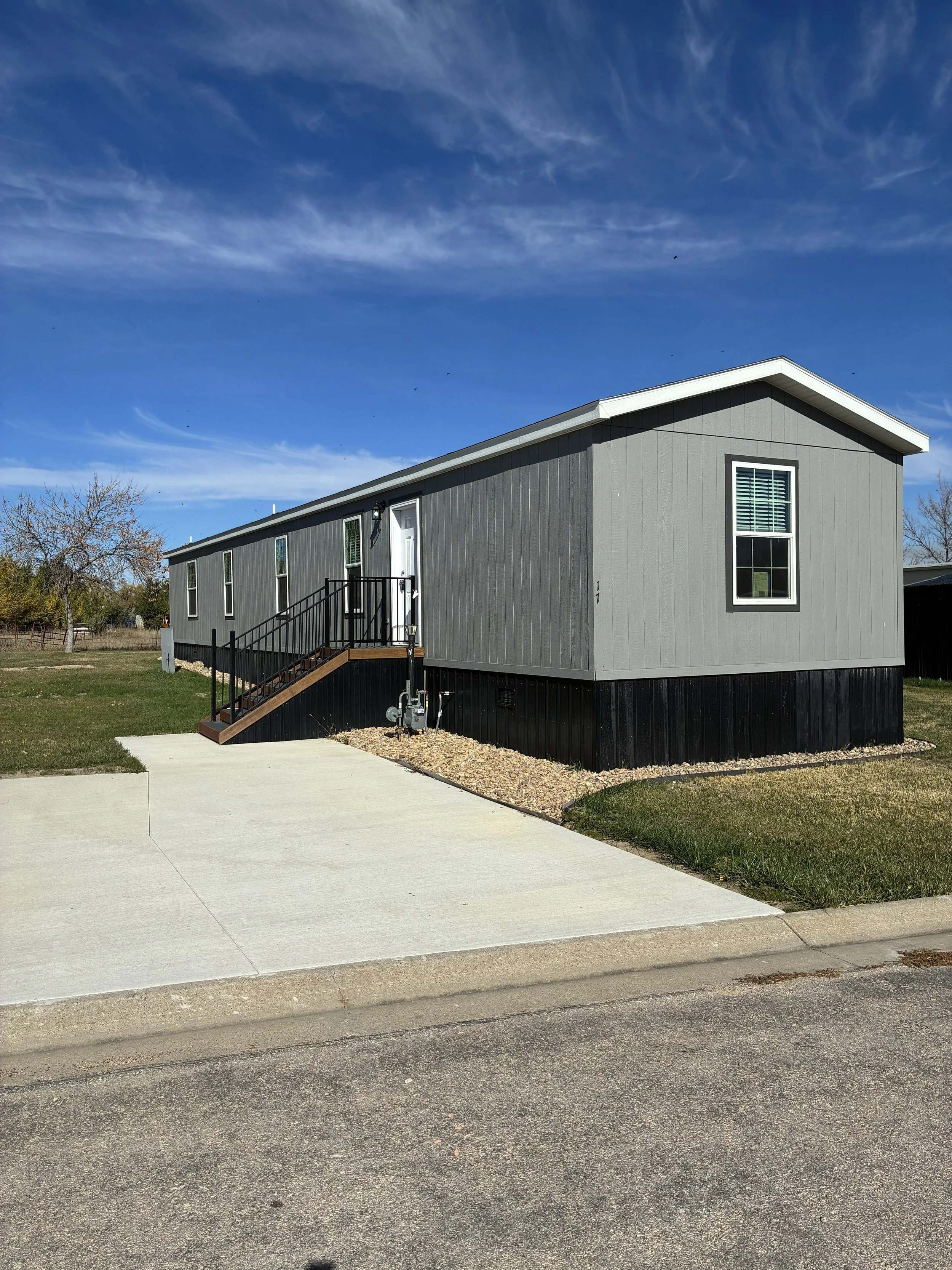 A gray manufactured home on a grassy lot with a concrete driveway, a small staircase, and a sign indicating it is for sale.