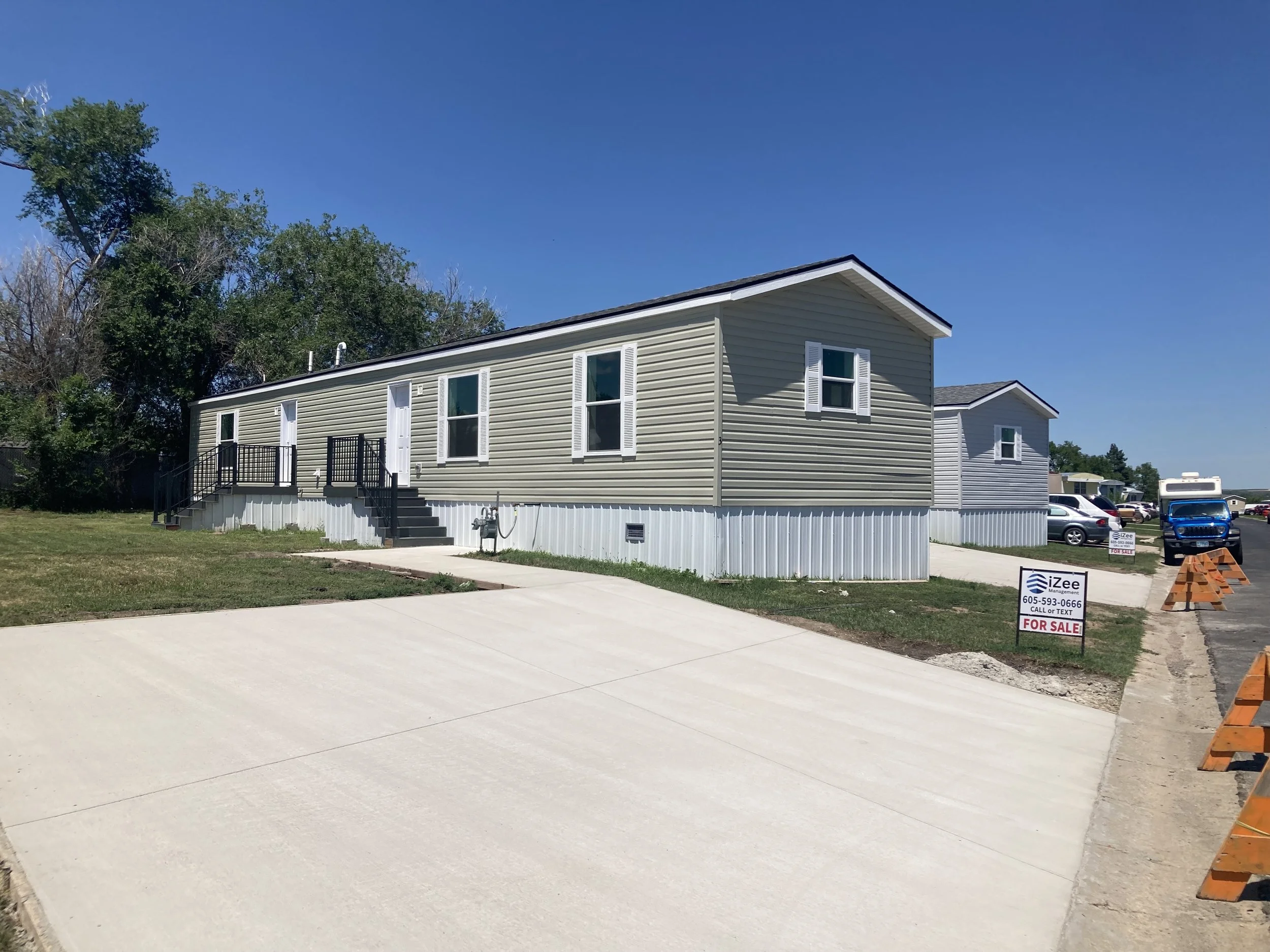 A mobile home with white siding and black stairs leading to the front doors, parked on a lot with some grass and a driveway, with a 