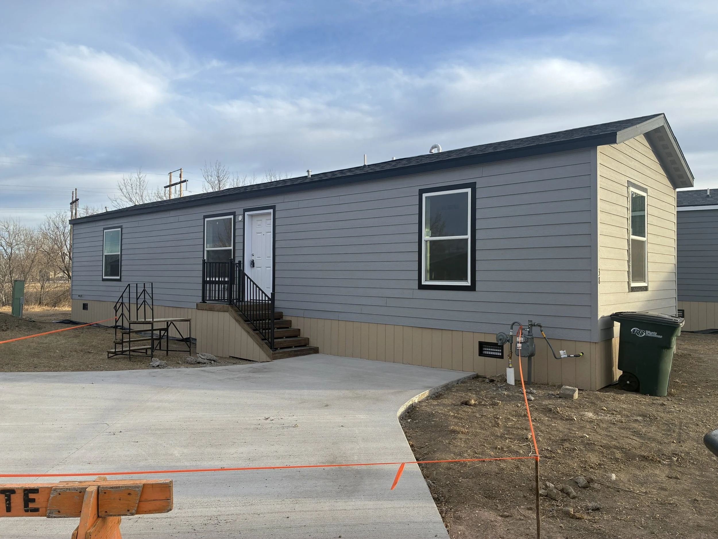 A gray manufactured home with white trim, four windows, and a small white staircase leading to the front door, set against a bright blue sky with a few clouds.