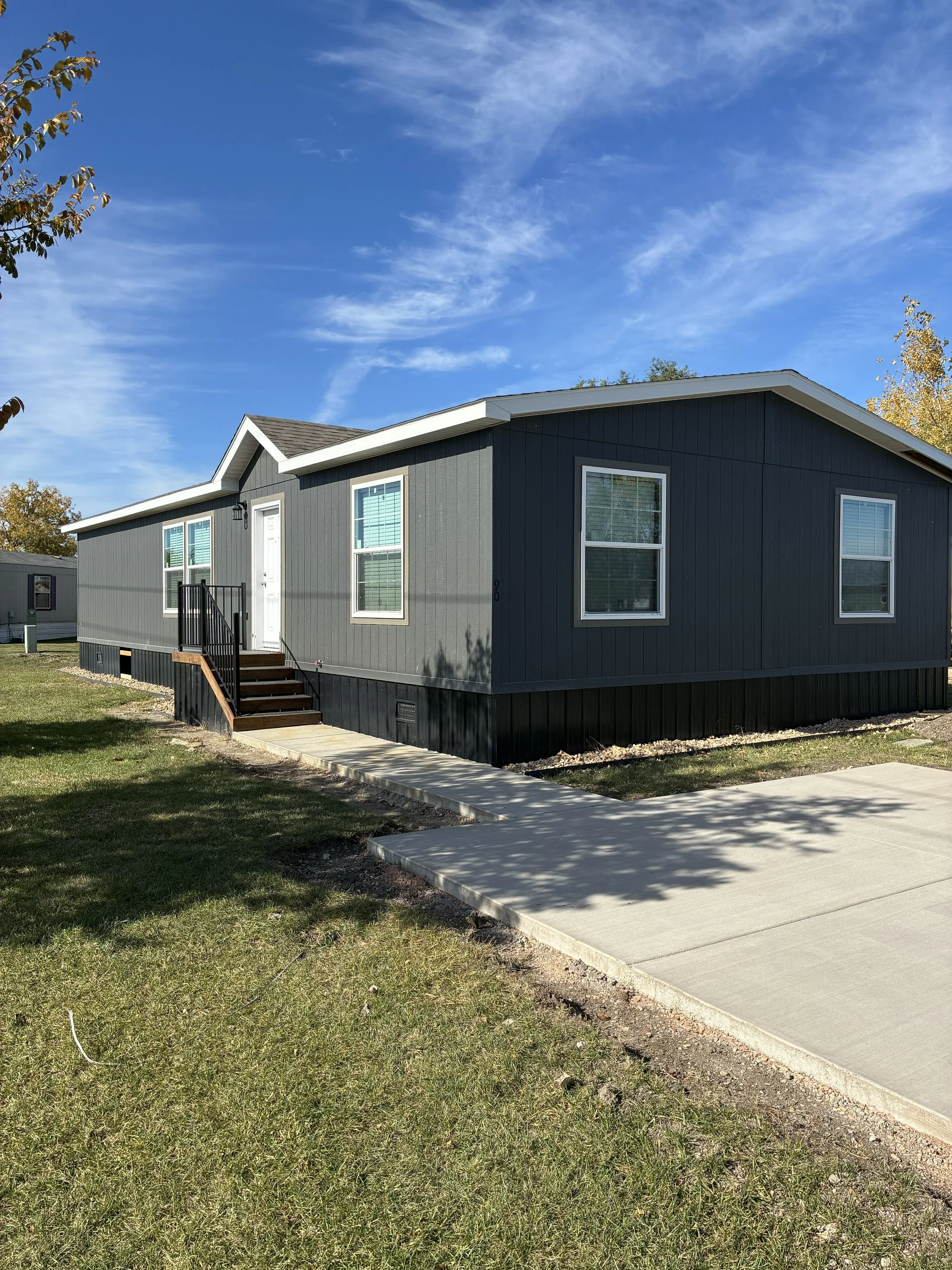 A black manufactured home on a grassy lot with a small set of stairs leading to a white door. Multiple windows on the front side and trees in the background under a blue sky.