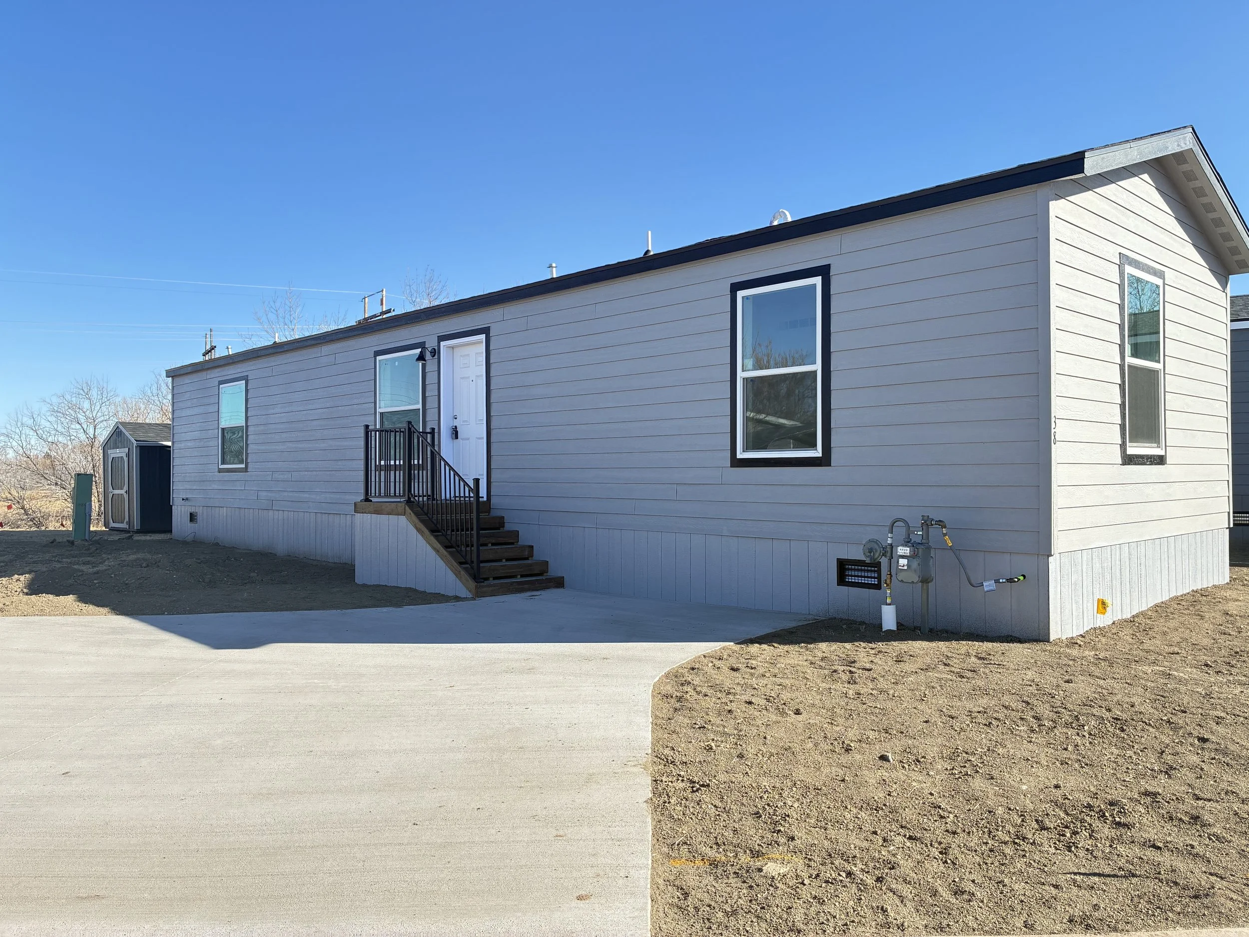 A gray manufactured home with white trim, four windows, and a small white staircase leading to the front door, set against a bright blue sky with a few clouds.