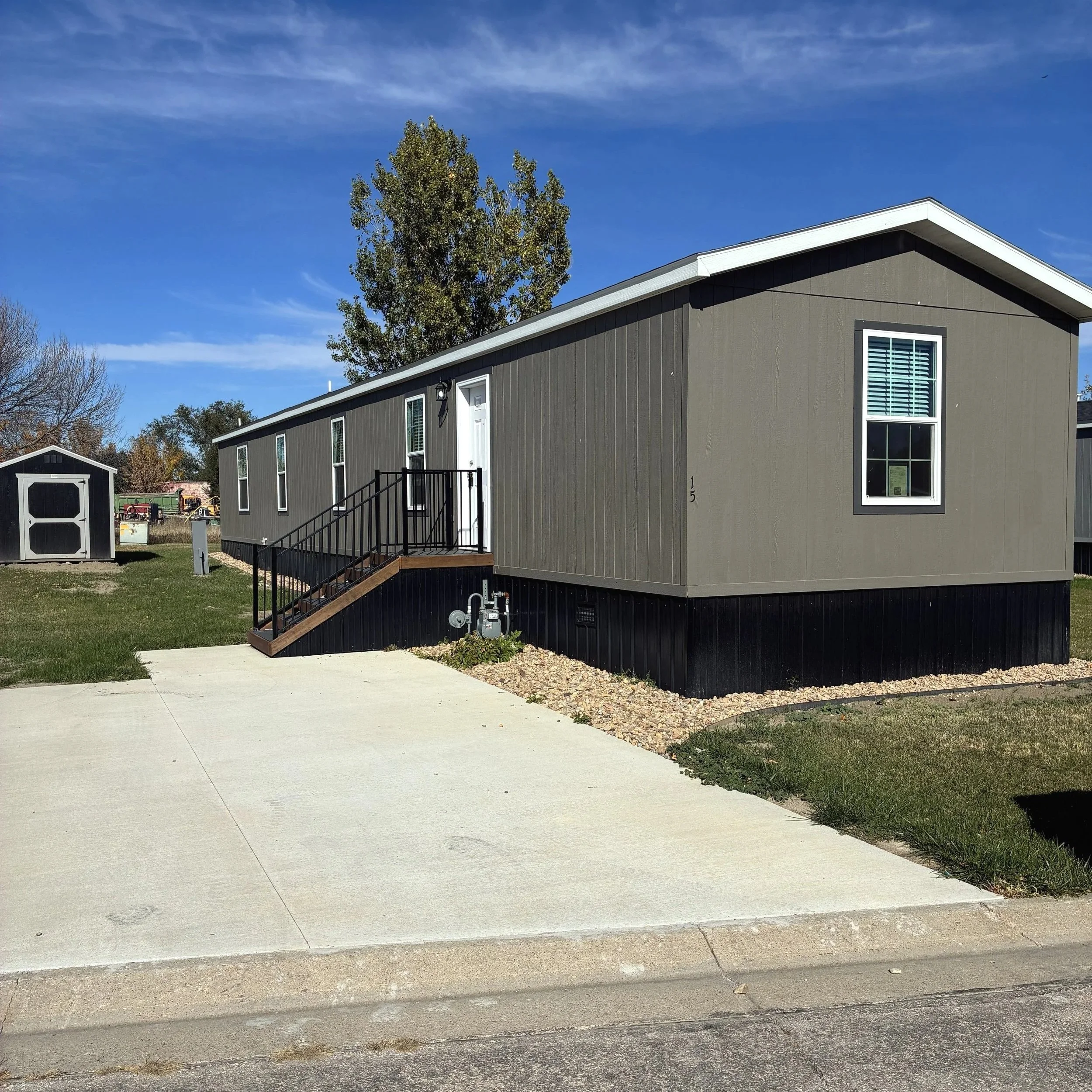 A manufactured home with a gray exterior, small front porch with stairs and black railing, and a driveway in front. There is a tree and blue sky in the background.