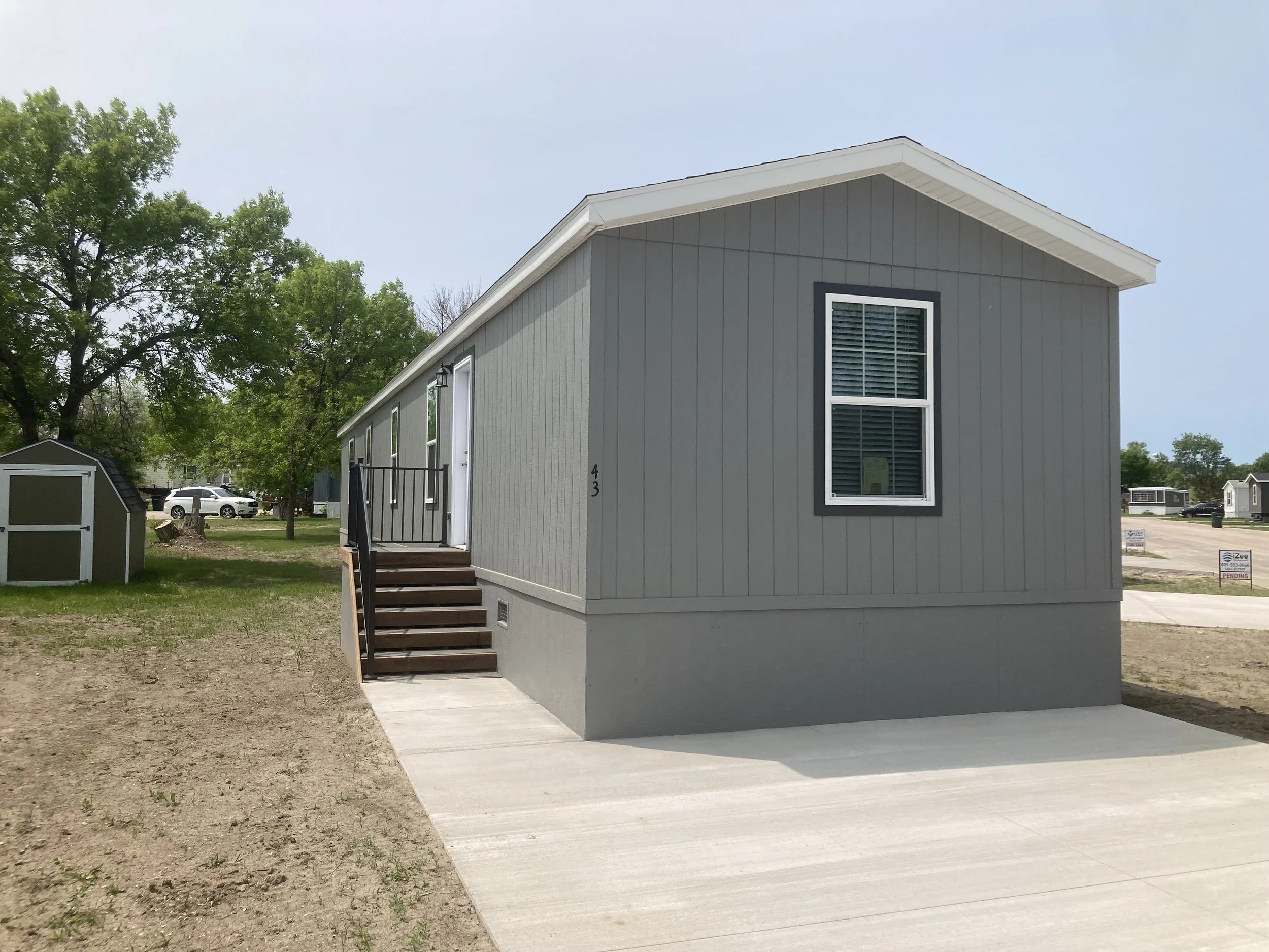 Newly constructed gray modular homes with small stoops, in a mobile home community with a paved driveway and signs for sale.