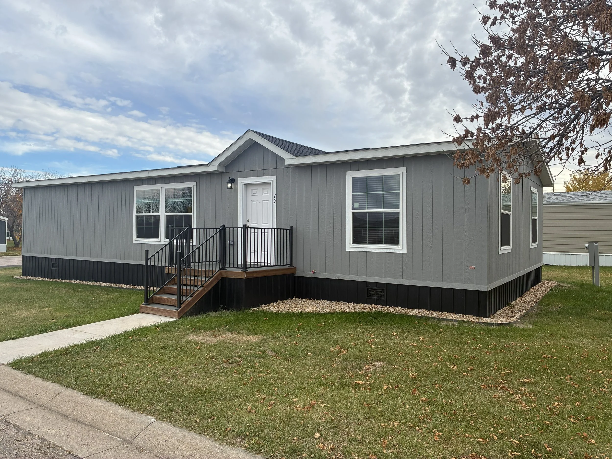 A small, gray manufactured home with white trim, a front door accessed by a set of wooden stairs with black railings, and four windows visible from the front, in a grassy area under a clear blue sky.