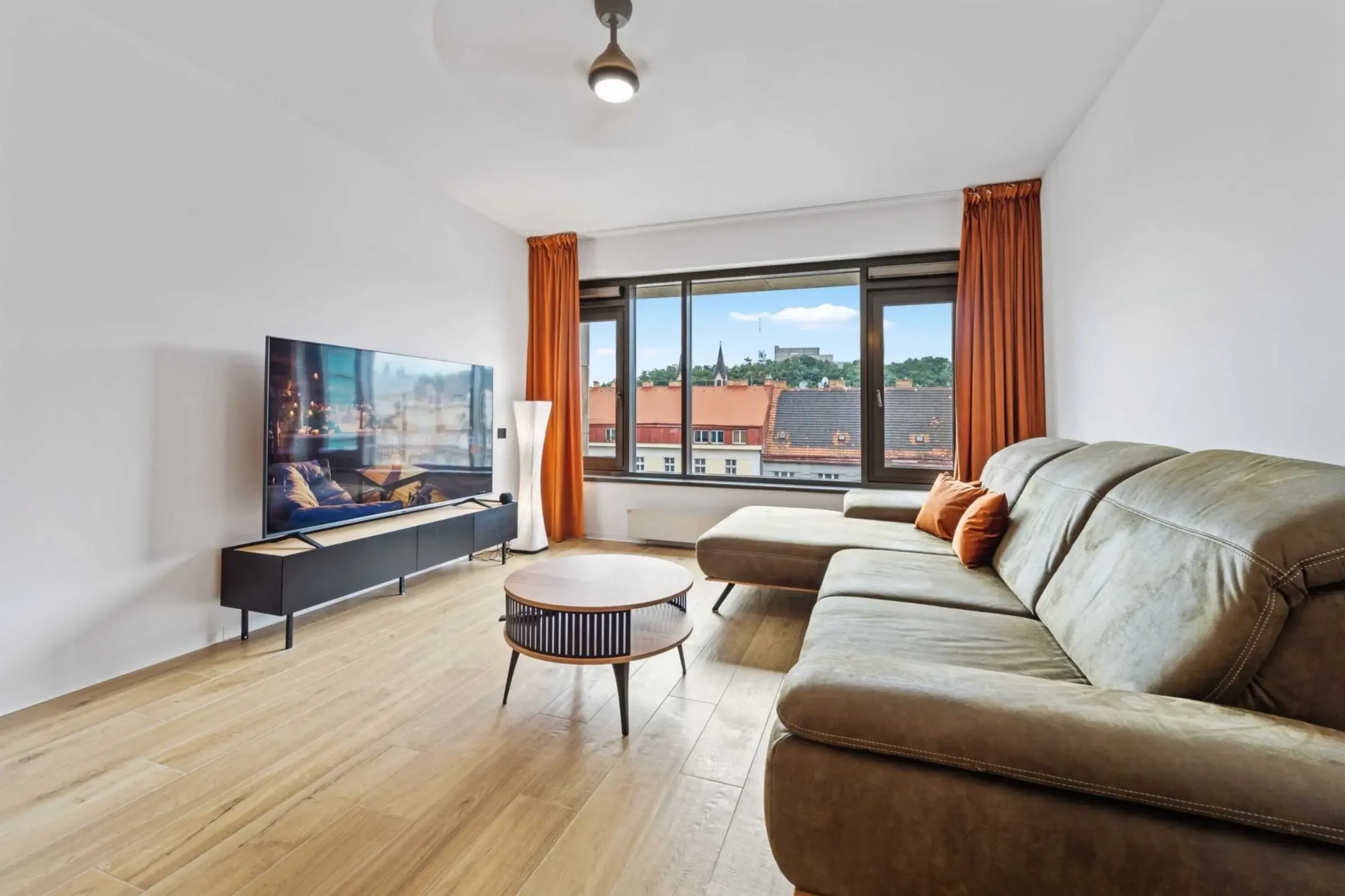 Modernes Wohnzimmer mit einem großen Sofa, einem runden Couchtisch, einem Flachbildfernseher auf einem schwarzen TV-Regal, einem weißen Lampenstandfuß, großen Fenstern mit orangefarbenen Vorhängen, und Blick auf ein Stadtbild mit roten Dächern und einem Kirchturm.