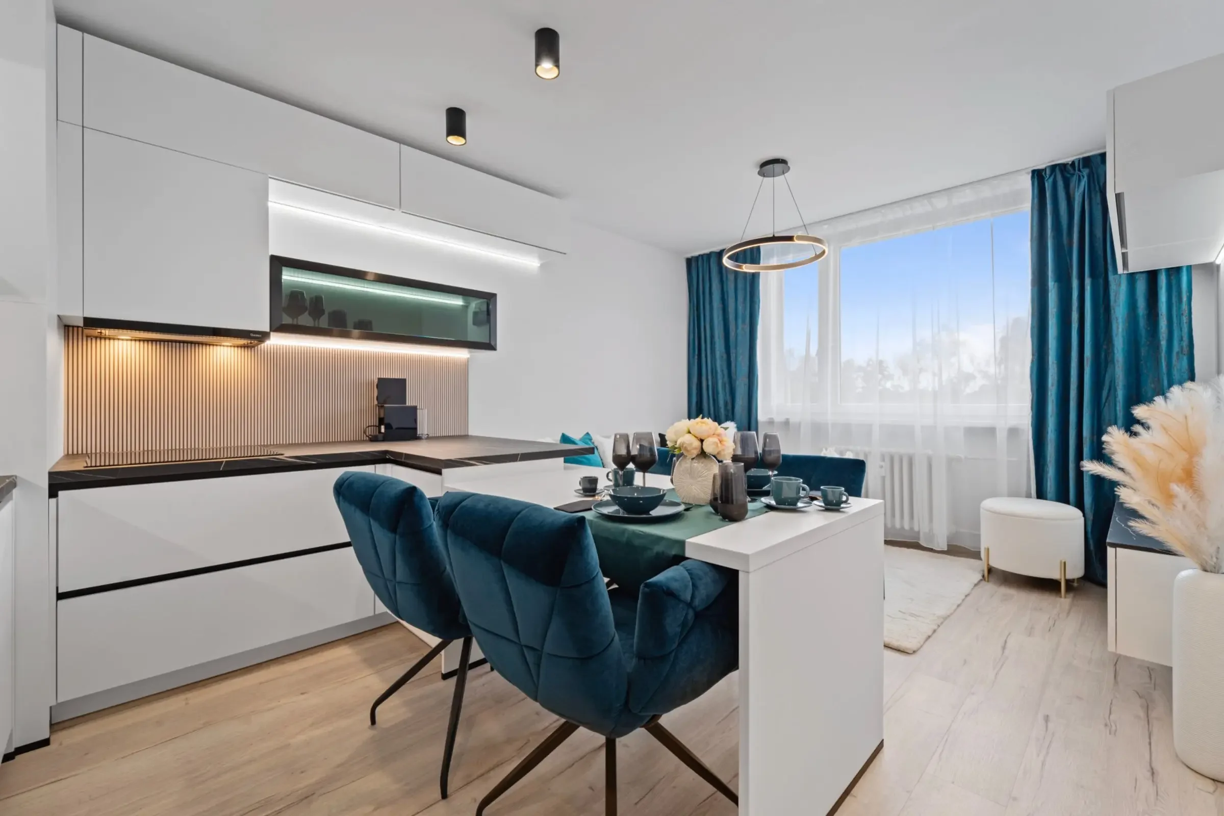 Moderne, offene Wohnung mit Essbereich, blauen Stühlen, weißen Möbeln und großen Fenstern mit blauen Vorhängen.