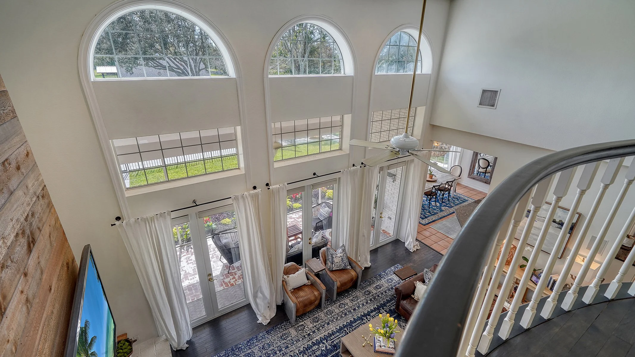 View of a living room with large arched windows and ceiling fan, white curtains, seating, and a glass sliding door leading to an outdoor patio.