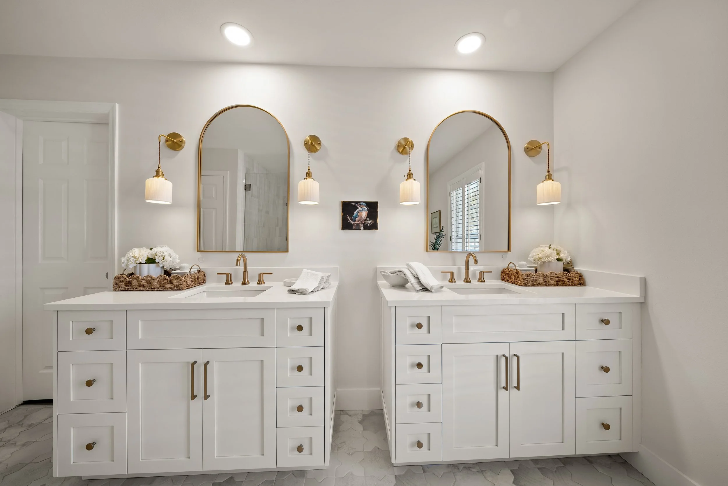 A modern bathroom featuring two white vanities with gold fixtures, each with an arched mirror, wall-mounted light fixtures, and decorative items such as flowers and towels. The bathroom has a neutral color scheme and a marble floor.