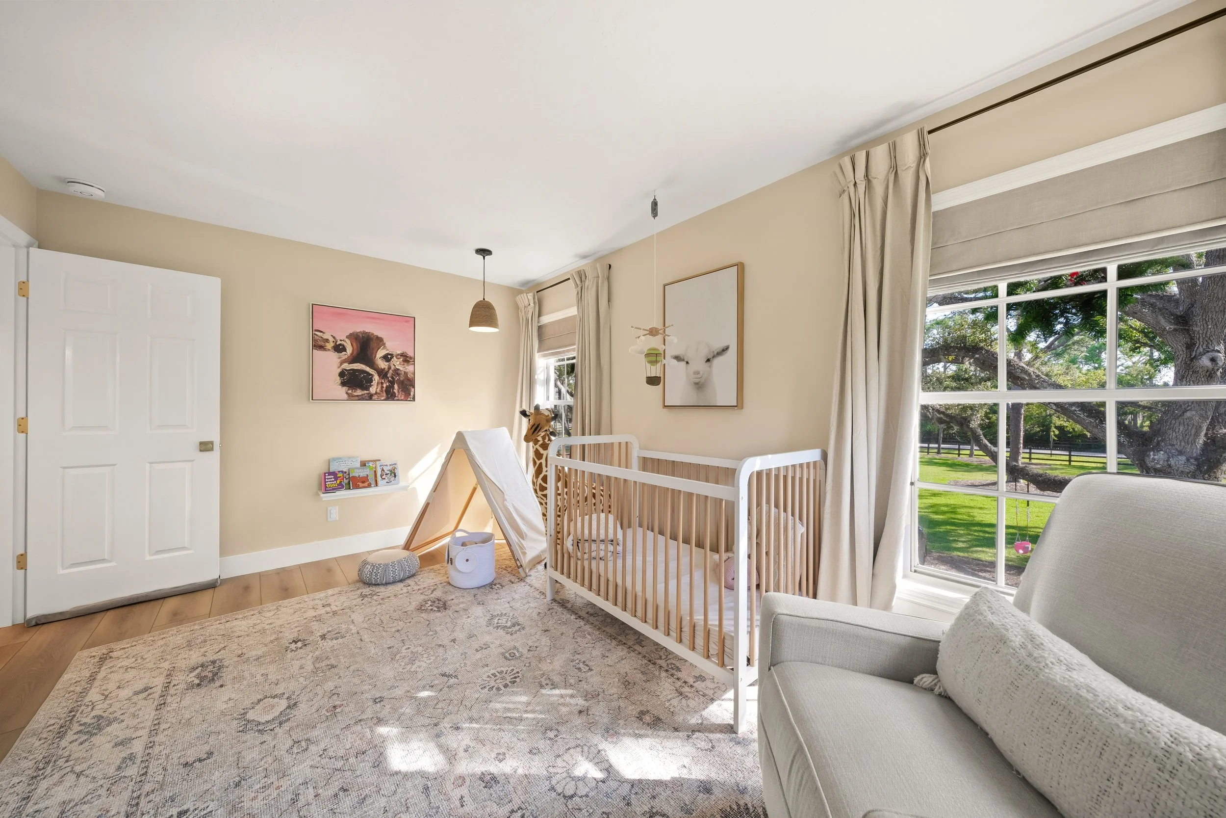 A cozy nursery room with a crib, a stuffed giraffe, artwork of animals, a teepee tent, books, and a large window showing a lush green yard outside.