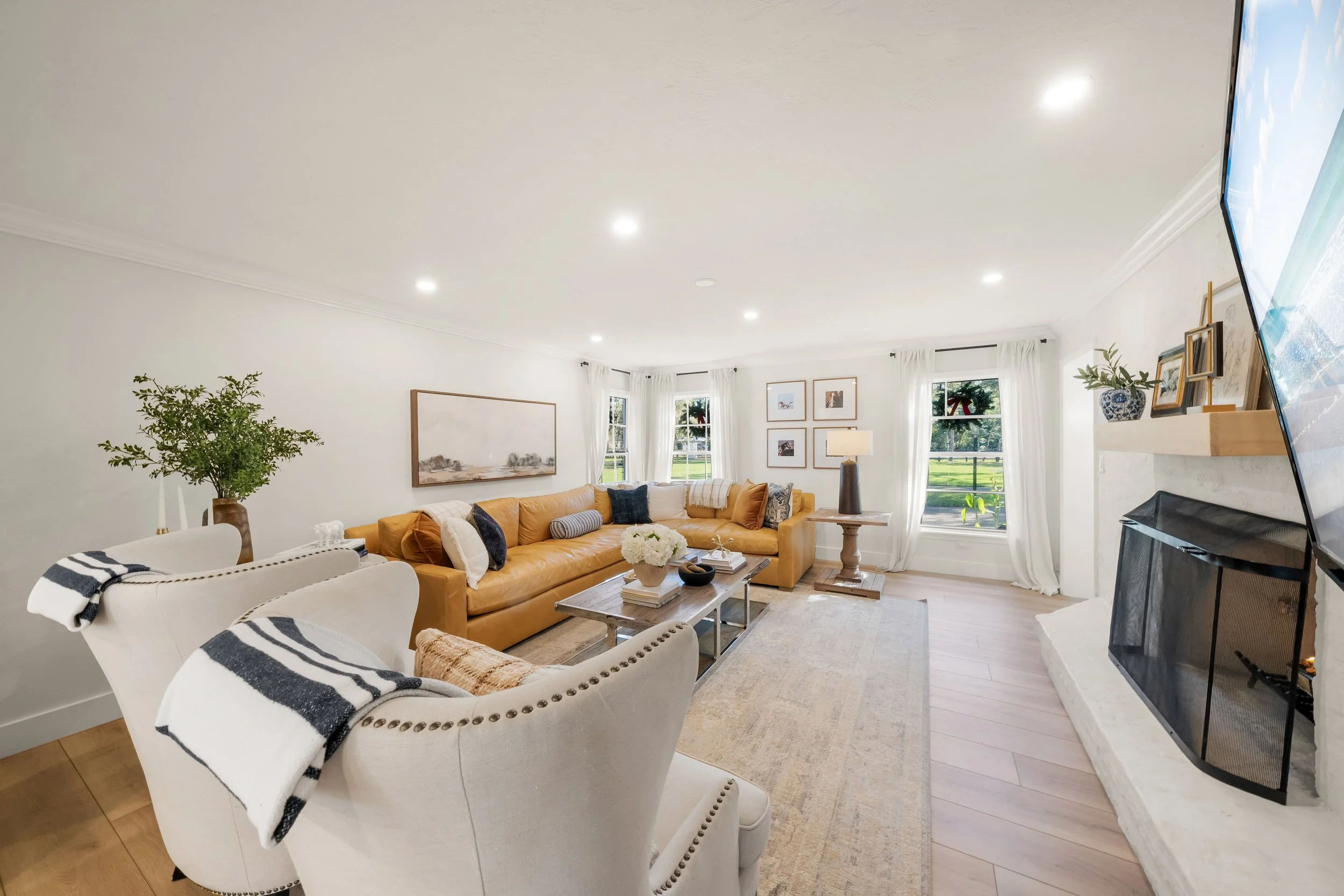 Bright living room with white walls, large windows with white curtains, tan leather sofa, white armchairs with black and white striped throws, a fireplace, and decorative items including framed art, family photos, and plants.