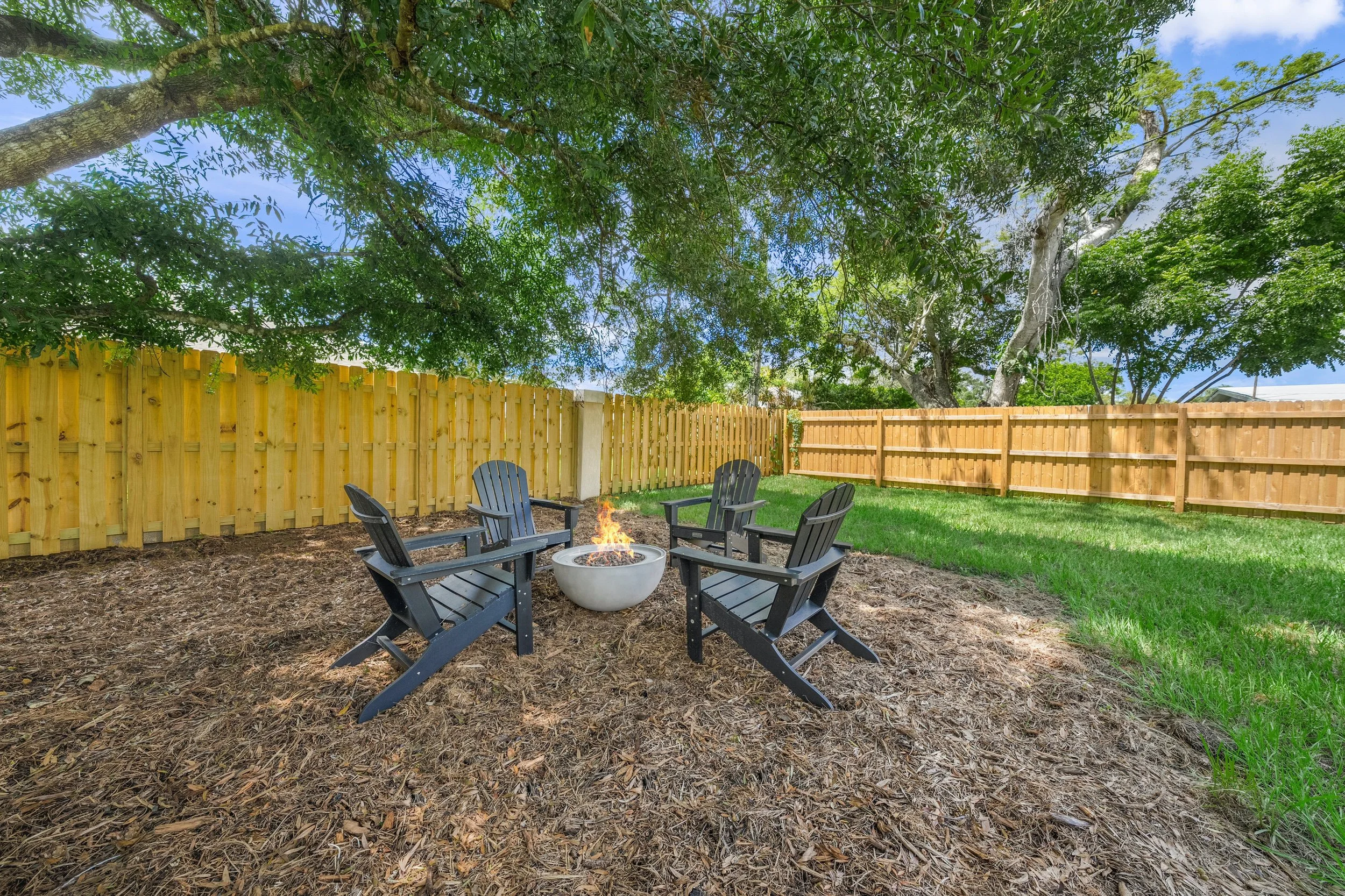 A backyard with four black Adirondack chairs arranged in a circle around a small fire pit with flames. The yard is surrounded by wooden fences, with large trees providing shade. There is a mix of mulch and grass on the ground.