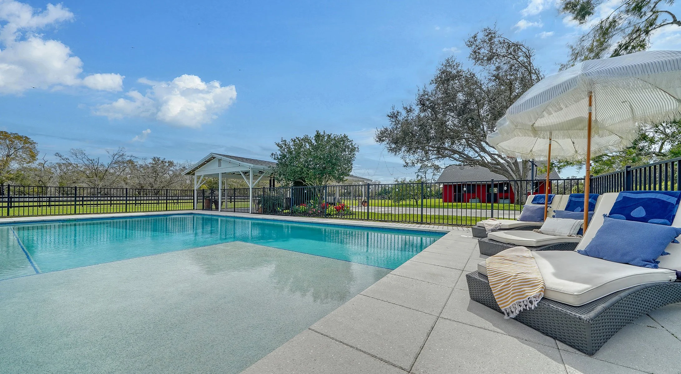 Outdoor swimming pool with lounge chairs and umbrellas, surrounded by a black fence, trees, and a red building in the background under a partly cloudy sky.
