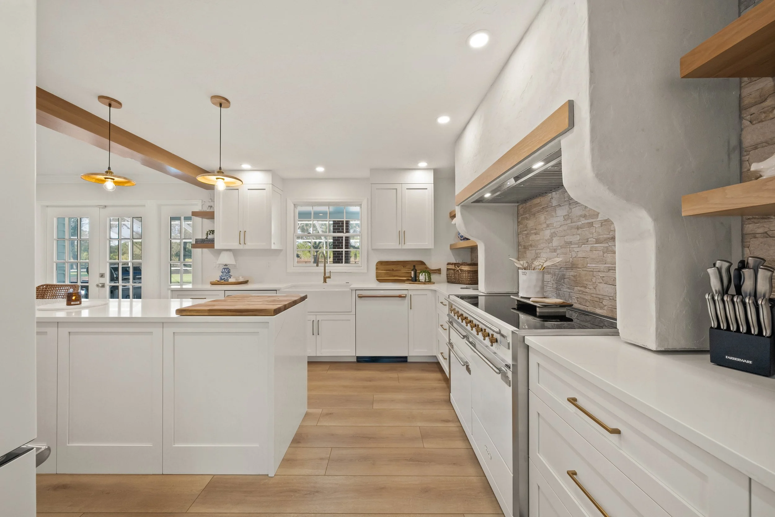 Modern white kitchen with wooden accents, pendant lights, and a central island, featuring a window and French door in the background.