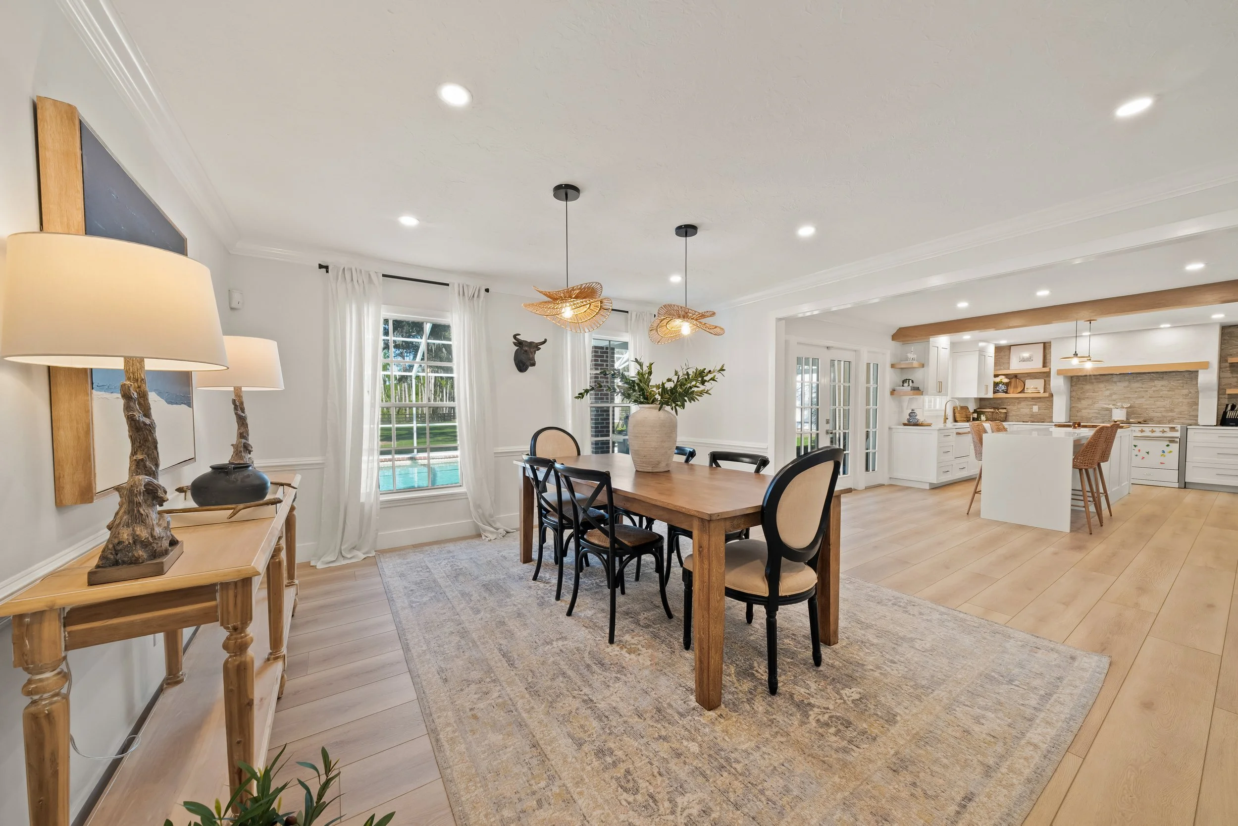 Bright open-plan dining and kitchen area with wooden dining table, six chairs, white cabinetry, and a kitchen island. Large windows with white curtains show a view of a backyard pool.