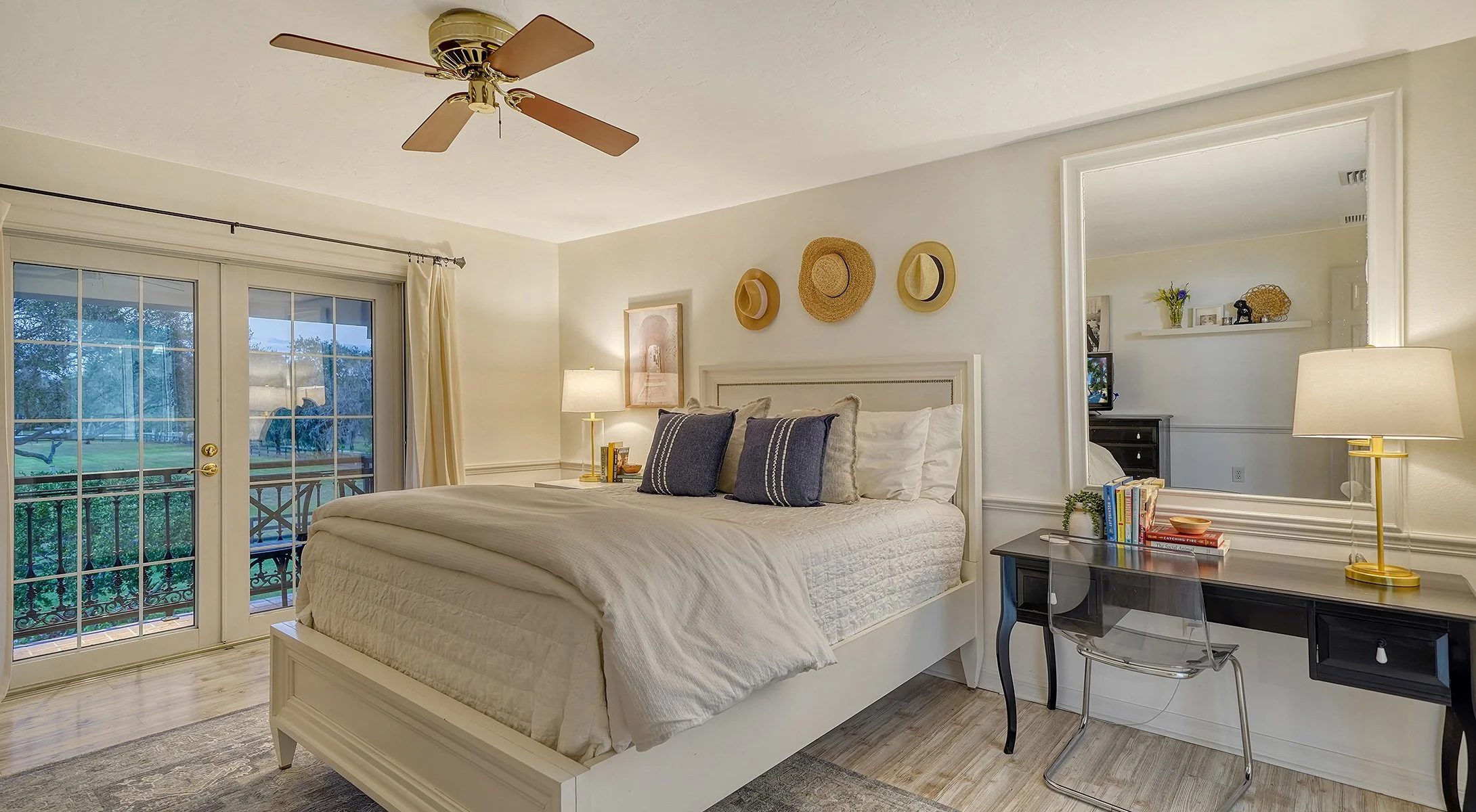 A cozy bedroom with a large window and glass sliding door leading to a balcony, a white bed with multiple pillows, a nightstand with a lamp, a black desk with a stack of books and a lamp, a large mirror, and decorative hats on the wall.