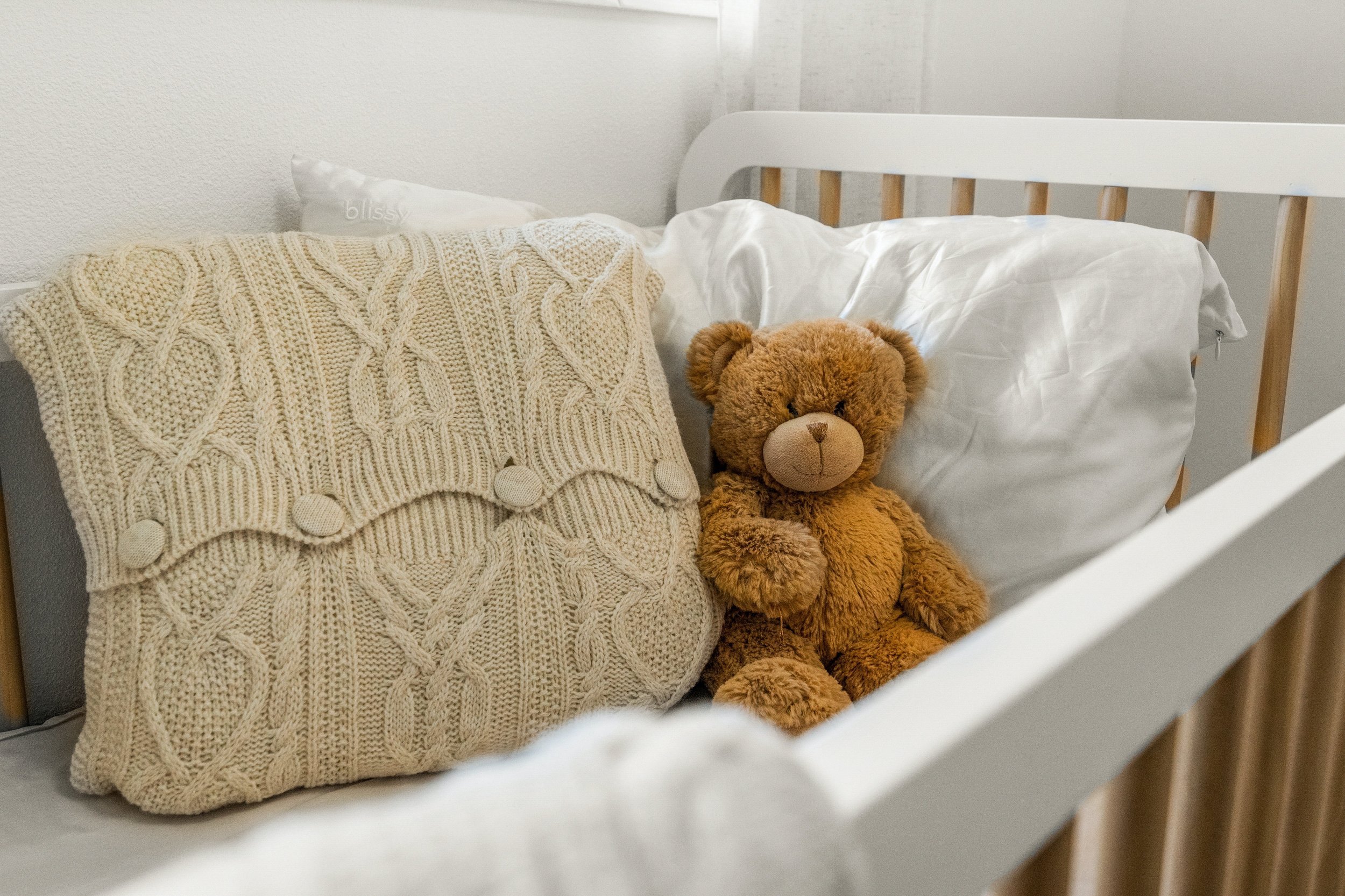 A teddy bear sitting on a bed next to a cream-colored knit pillow with pom-pom embellishments.