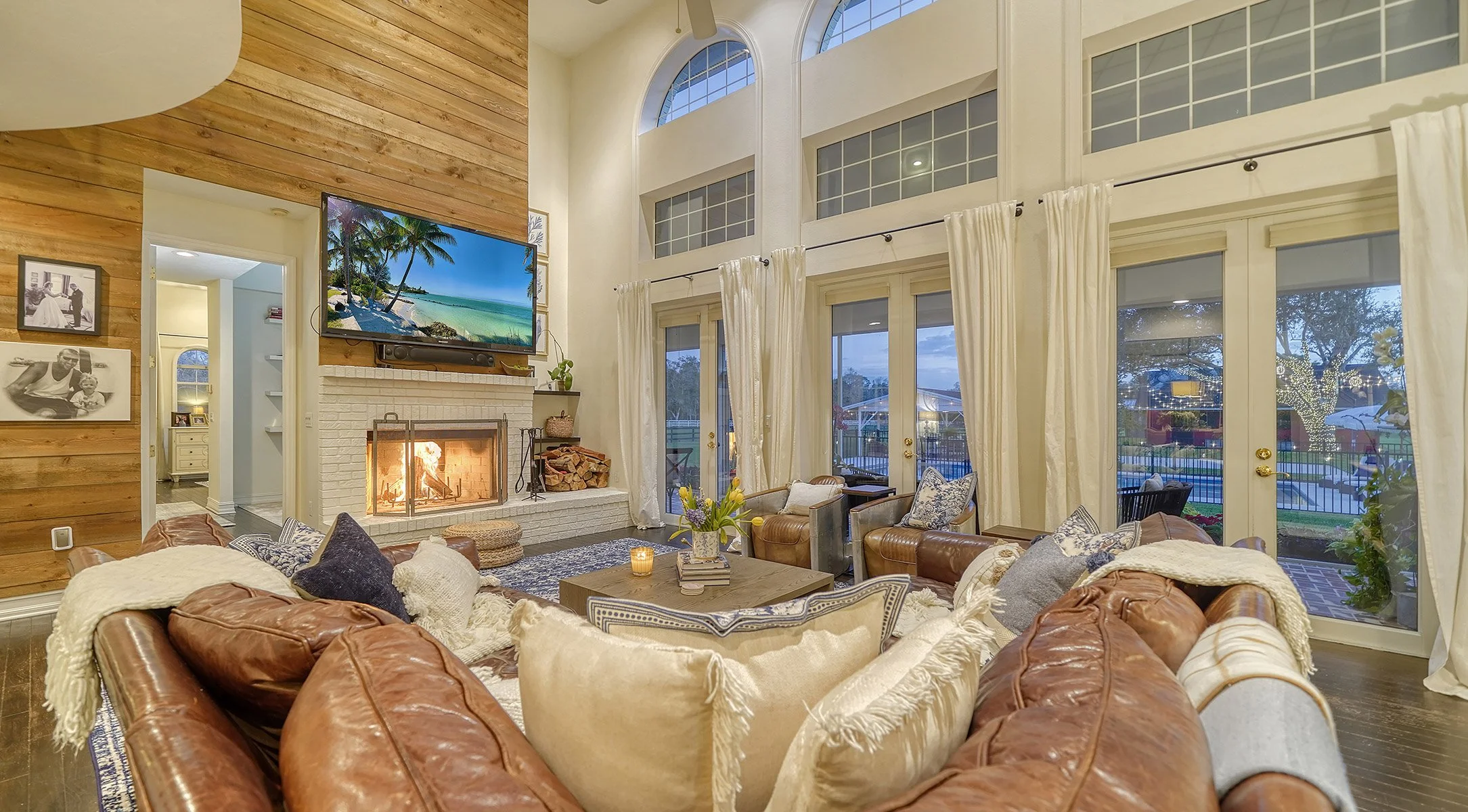 Living room with leather sofas, a central coffee table, a fireplace, and large windows with curtains. A wall-mounted TV shows a beach scene. The room has a high ceiling and wood-paneled wall, with a view of a backyard with trees and string lights.