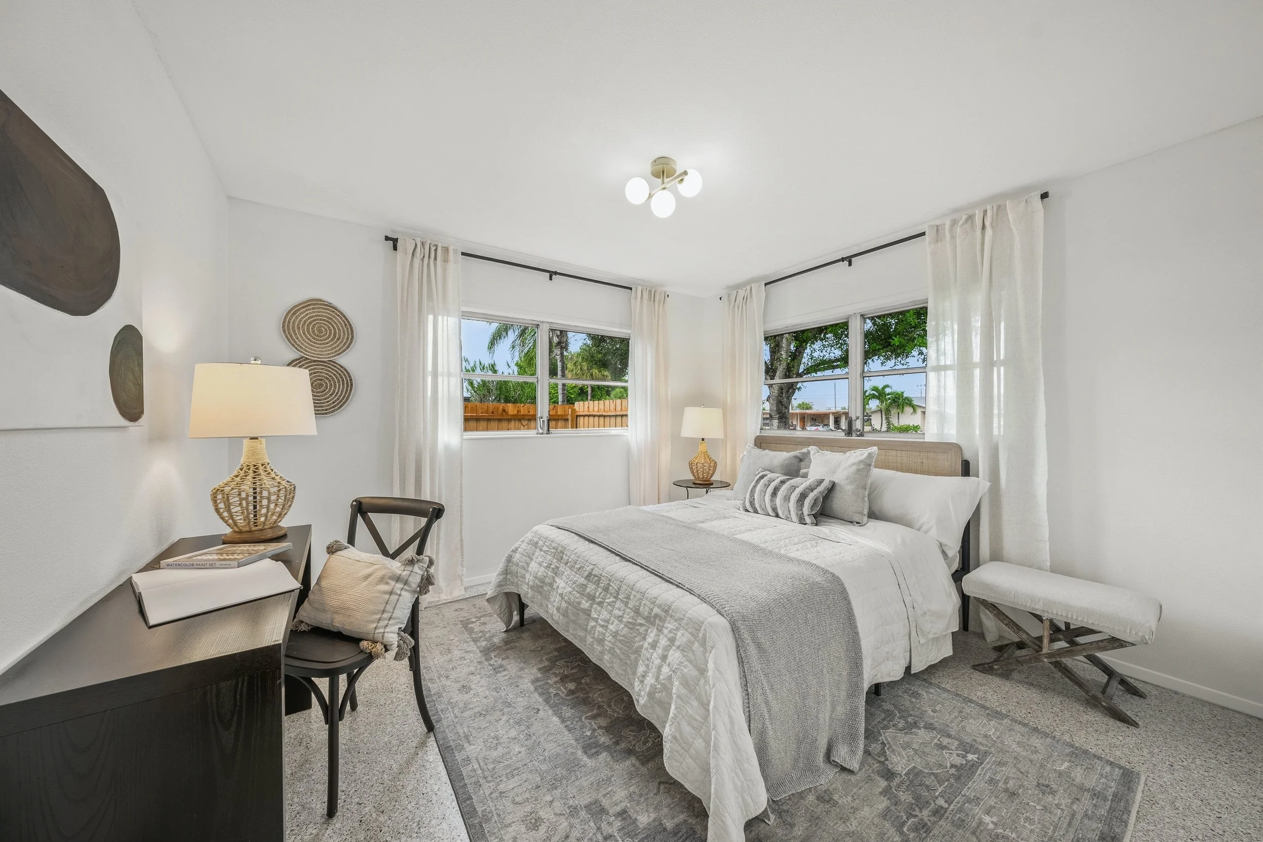 A bedroom with a bed, bedside lamps, windows with curtains, artwork on the wall, a chair with a pillow, and a bench at the foot of the bed, during daytime.