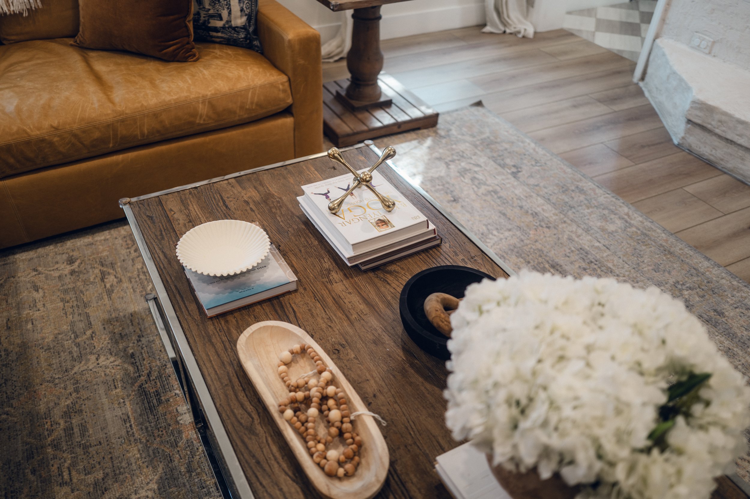 A wooden coffee table with books, a decorative metal cross, a black dish, a wooden tray with beads, and a large bouquet of white flowers. A tan leather sofa and a wooden side table are in the background.