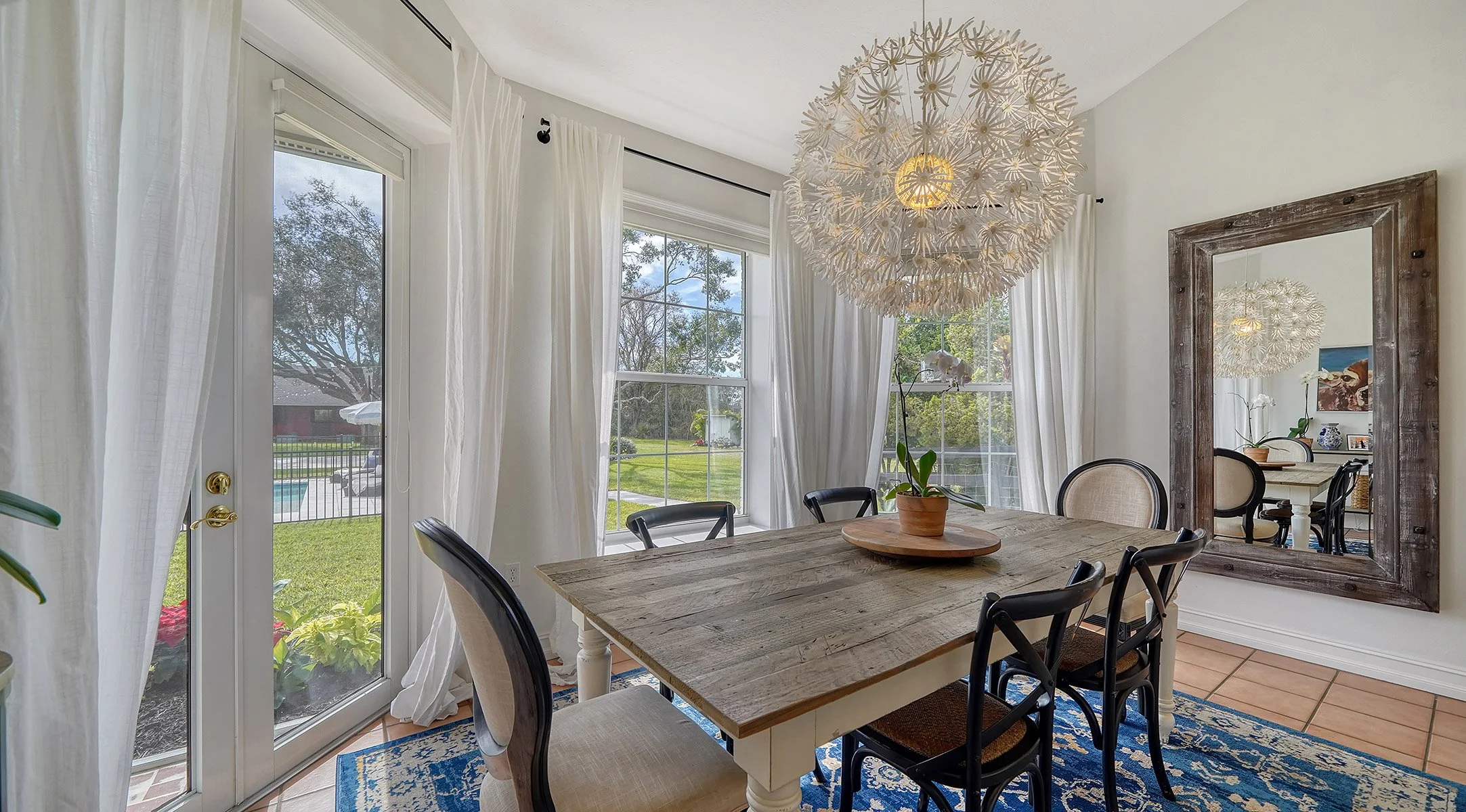 A dining room with a wooden table, six chairs, a large mirror, and a pendant light fixture. Large windows with white curtains show a yard with trees and a pool outside.