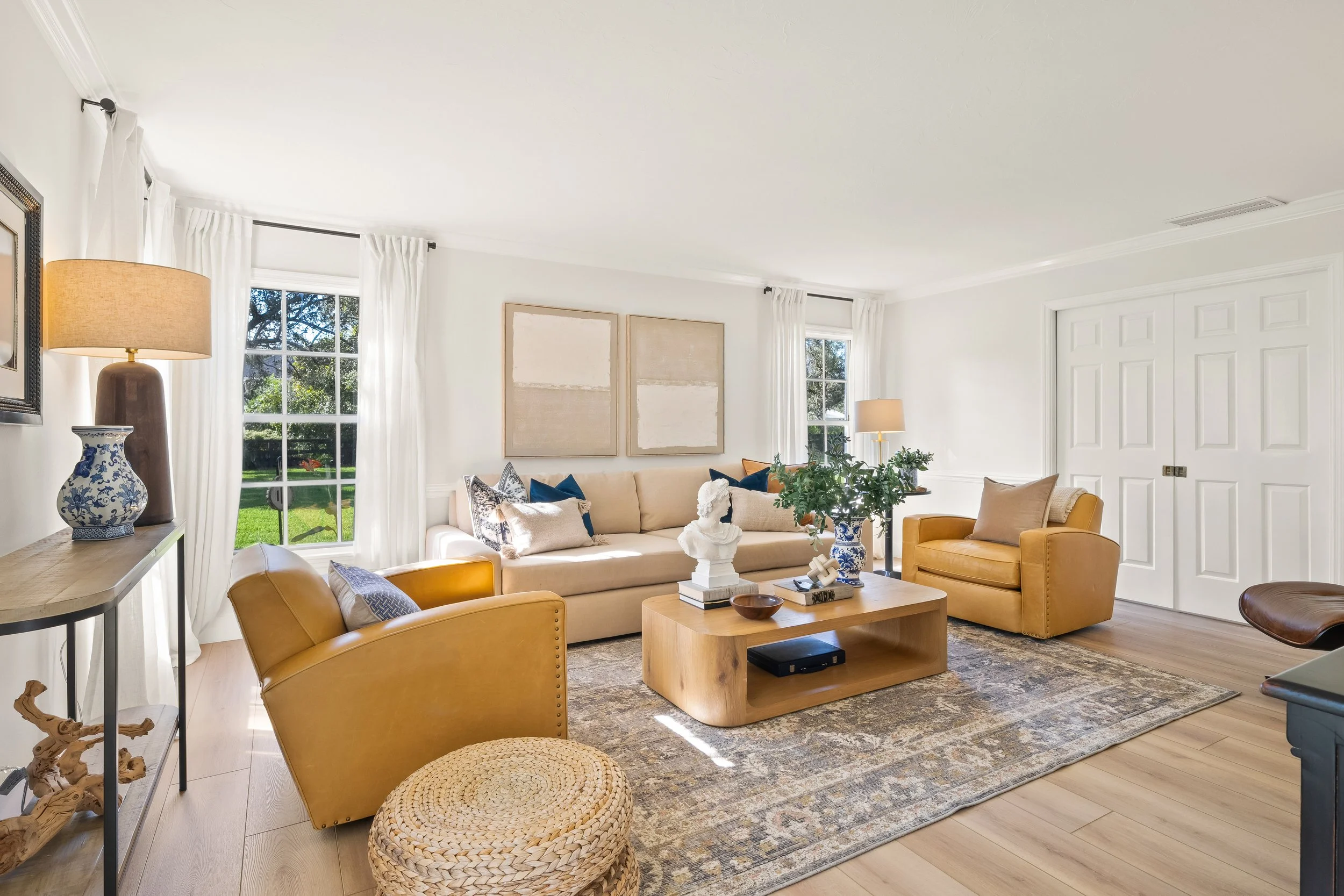 Bright living room with beige sofa, yellow armchairs, wooden coffee table, artworks, lamps, and large windows with white curtains, overlooking a green yard.