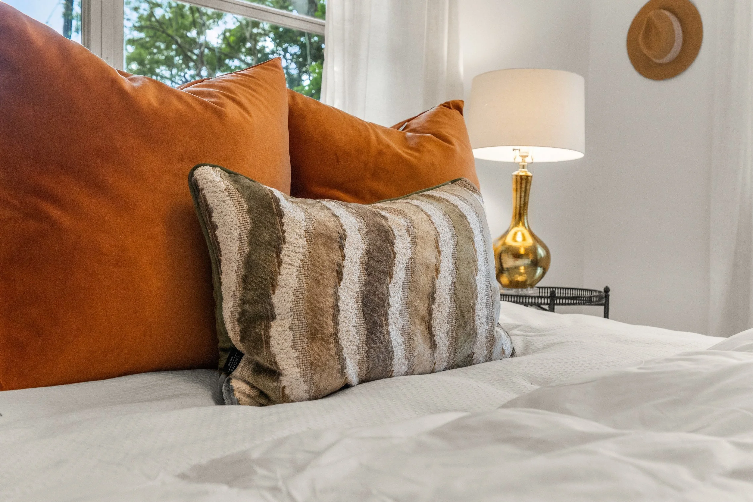 A bed with orange velvet pillows, a decorative striped pillow, a white bedsheet, a gold table lamp, and a window showing green trees outside.