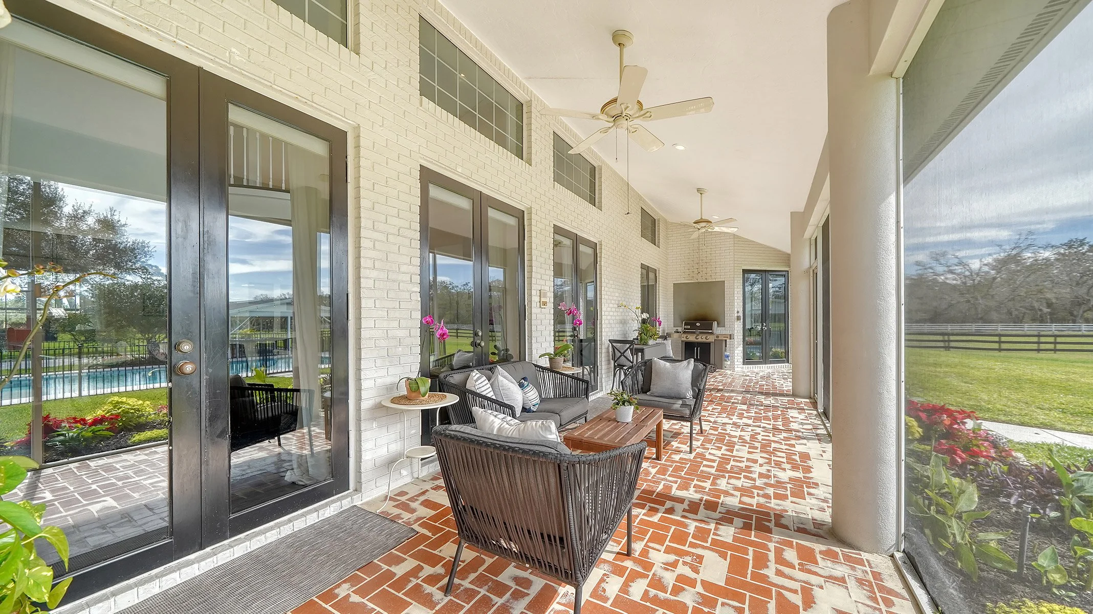 Outdoor screened porch with black wicker furniture, including a sofa and chairs with cushions, a brick-patterned floor, plants, a barbecue grill, and a view of a backyard with grass and trees.