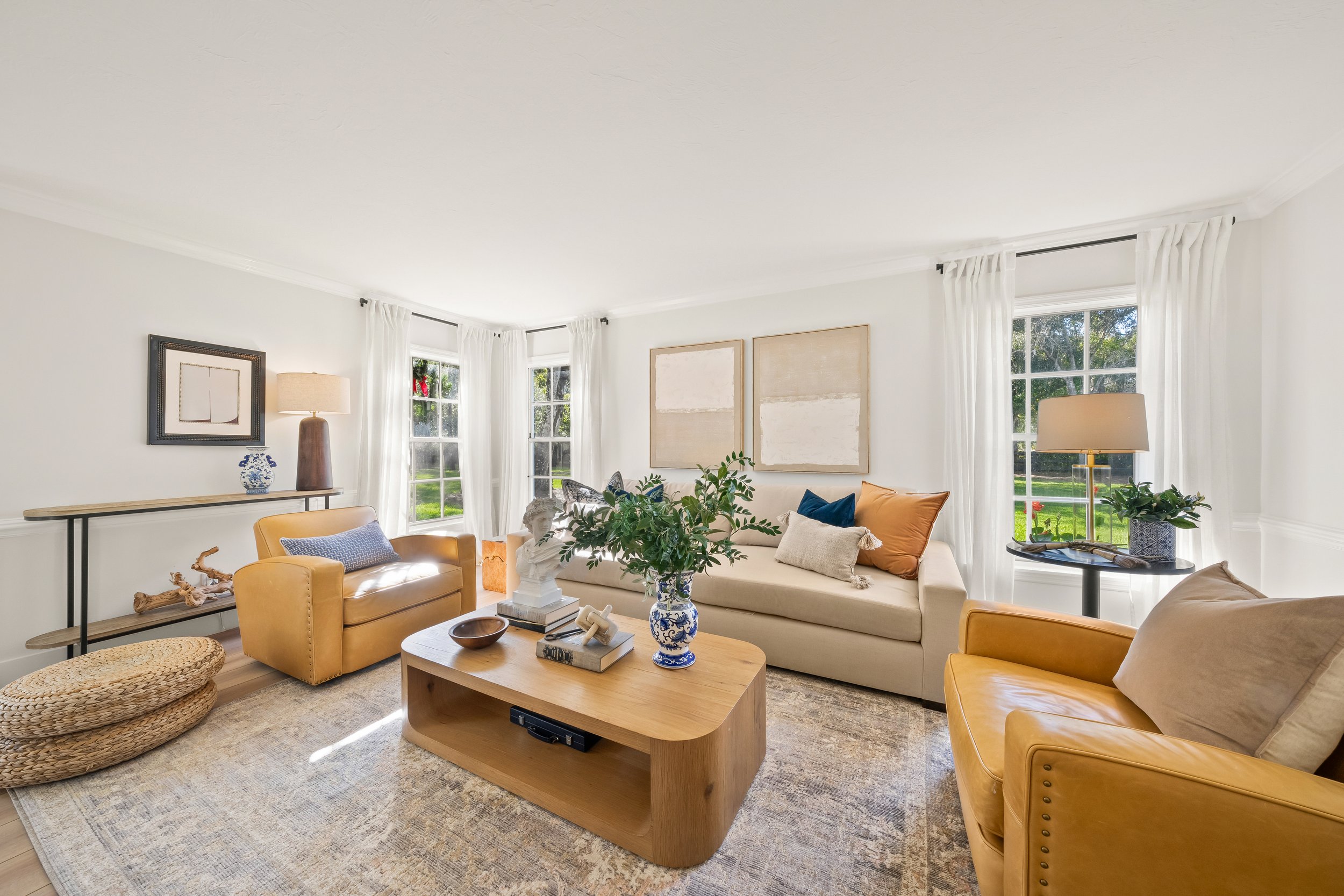 Bright living room with cream walls, white curtains, a beige sofa with colorful pillows, yellow armchairs, a wooden coffee table with decorative items, and large windows with greenery outside.
