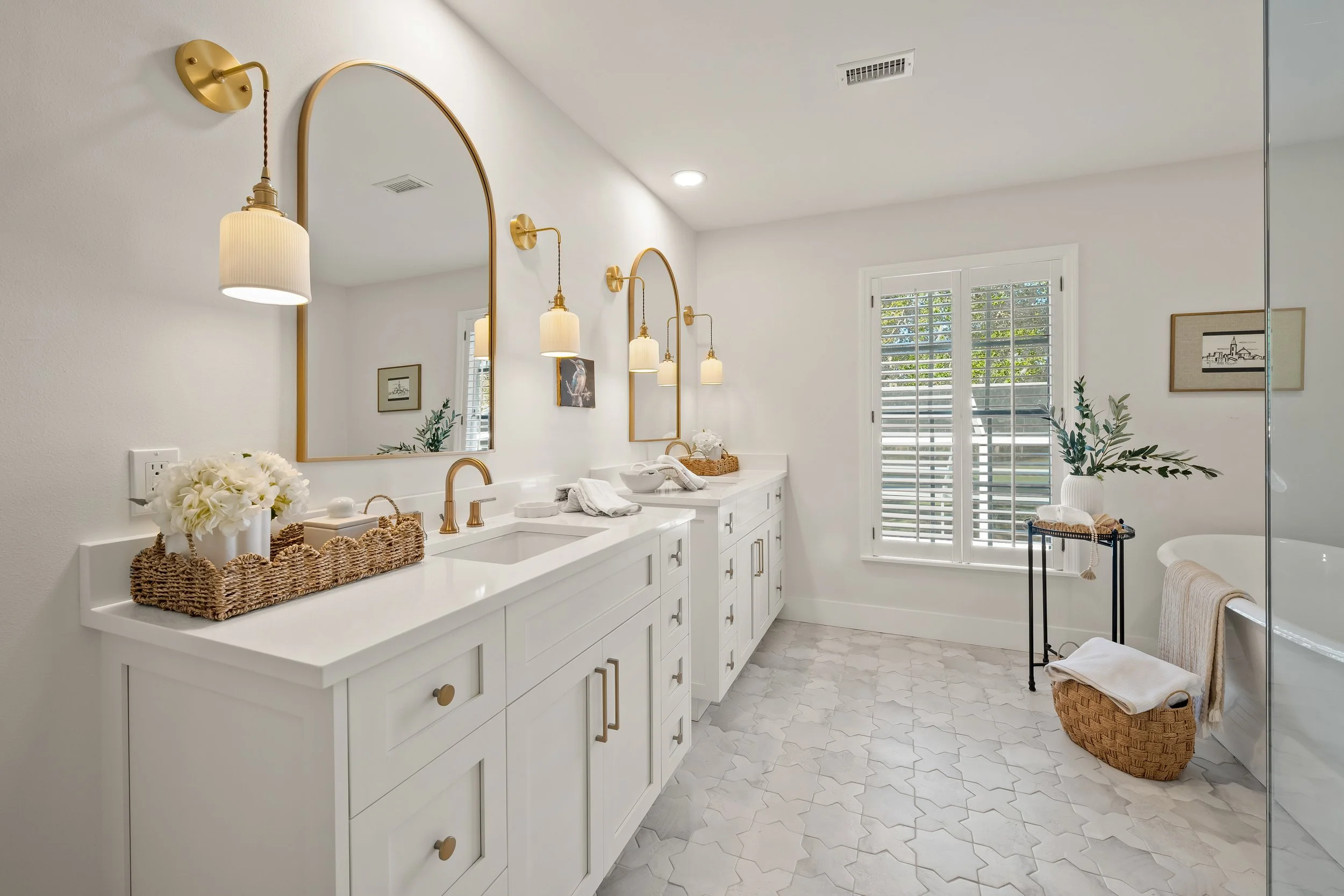 Bright white bathroom with double vanity, gold fixtures, large mirror, and natural light through a window with shutters. Decor includes flowers, framed art, and a basket of towels.