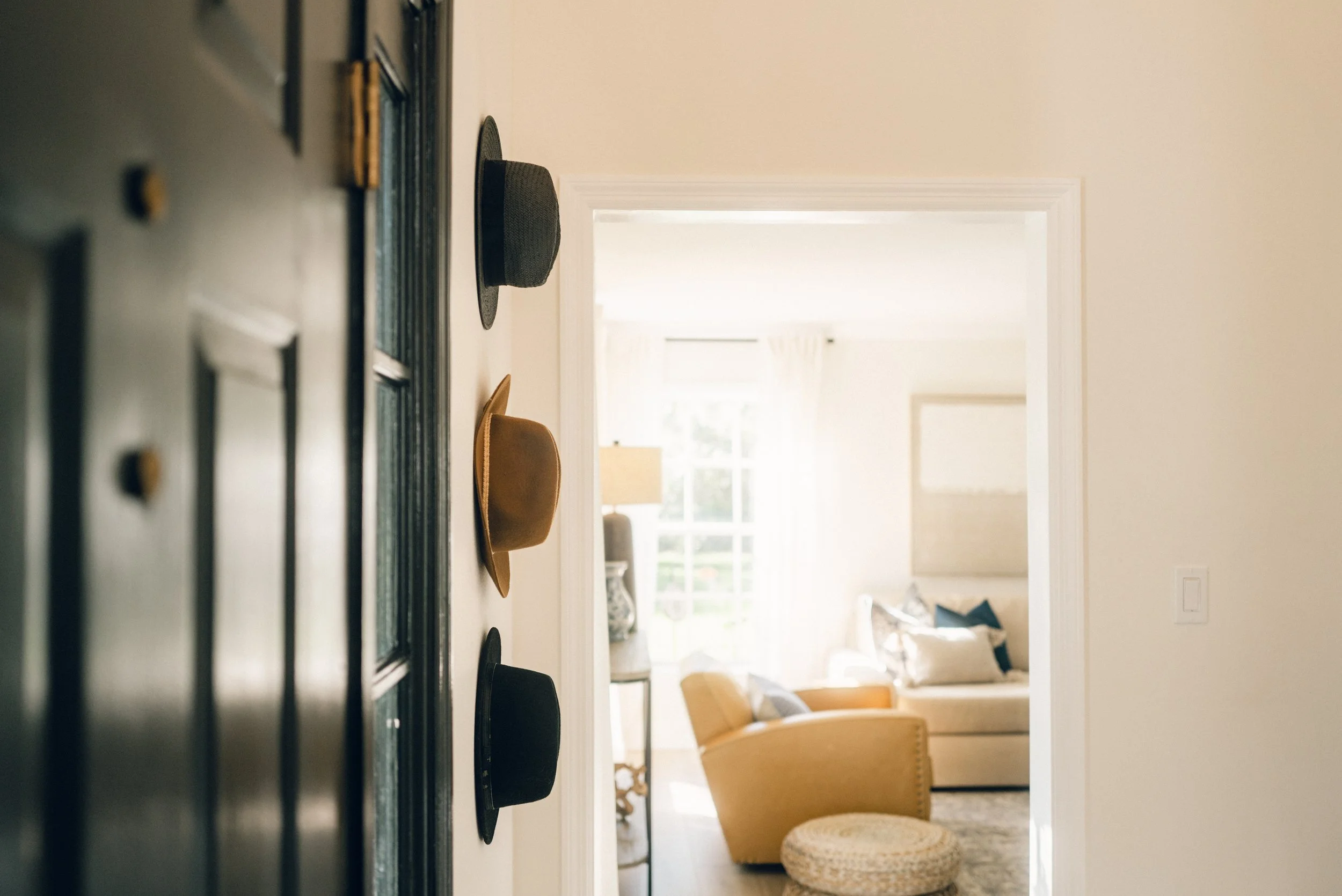 Hat hooks on a white wall with three hats, leading into a bright living room with a yellow armchair, sofa, and window.