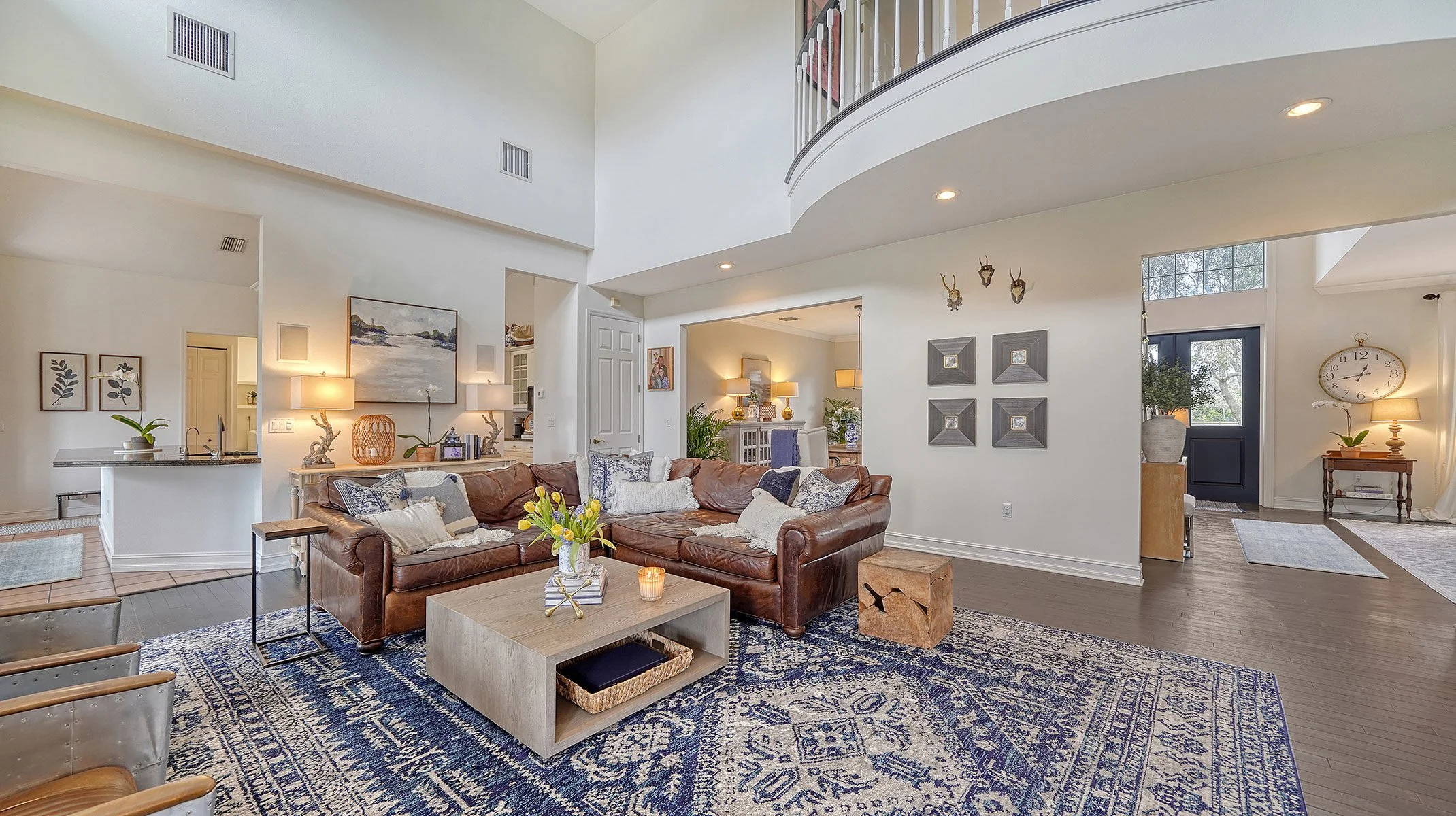Spacious living room with a brown leather sectional sofa, navy and white patterned rug, and modern decor, including wall art, lamps, and a glass vase with yellow flowers.