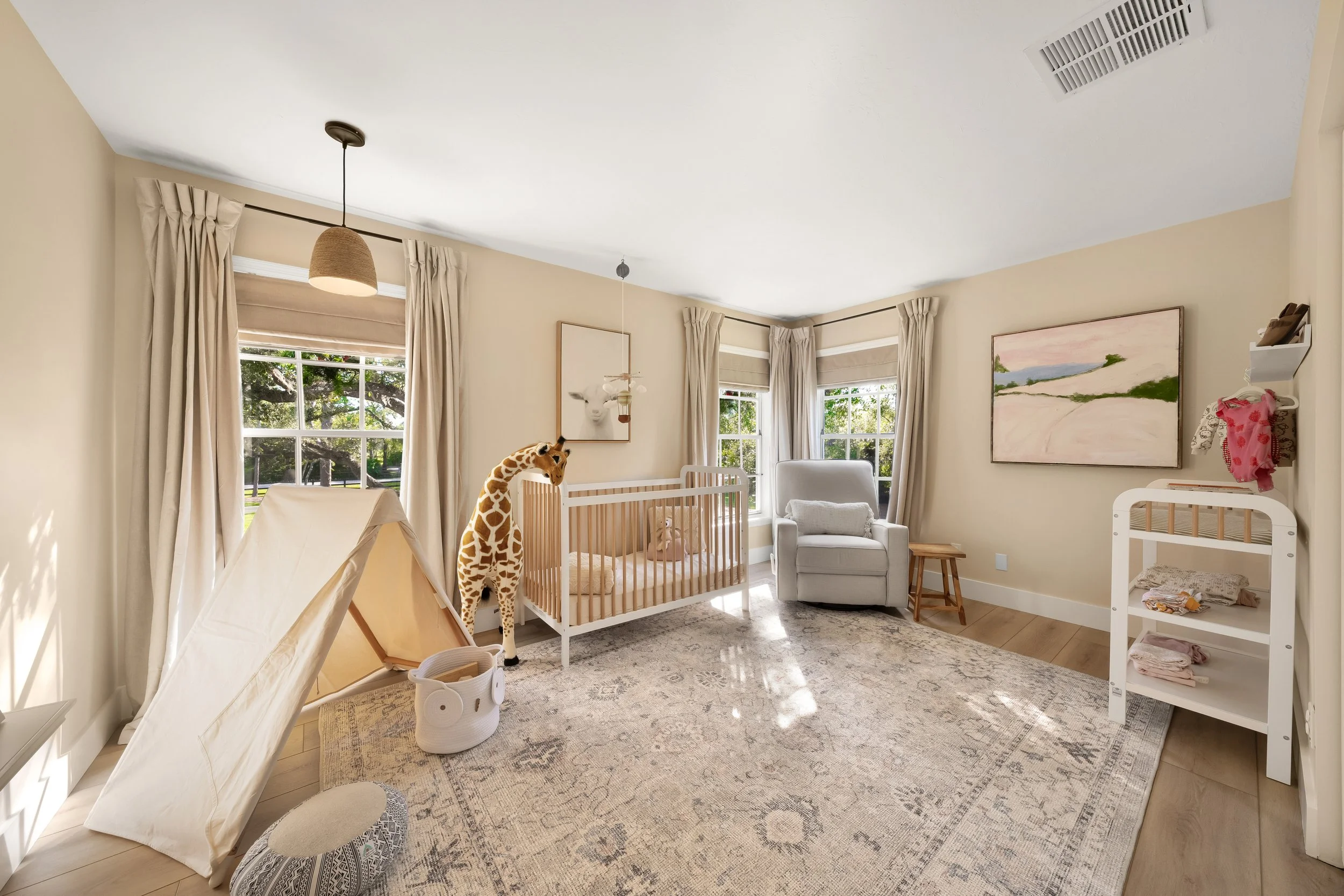 Child's nursery with a crib, stuffed giraffe, tent, armchair near windows, and artwork on the walls.